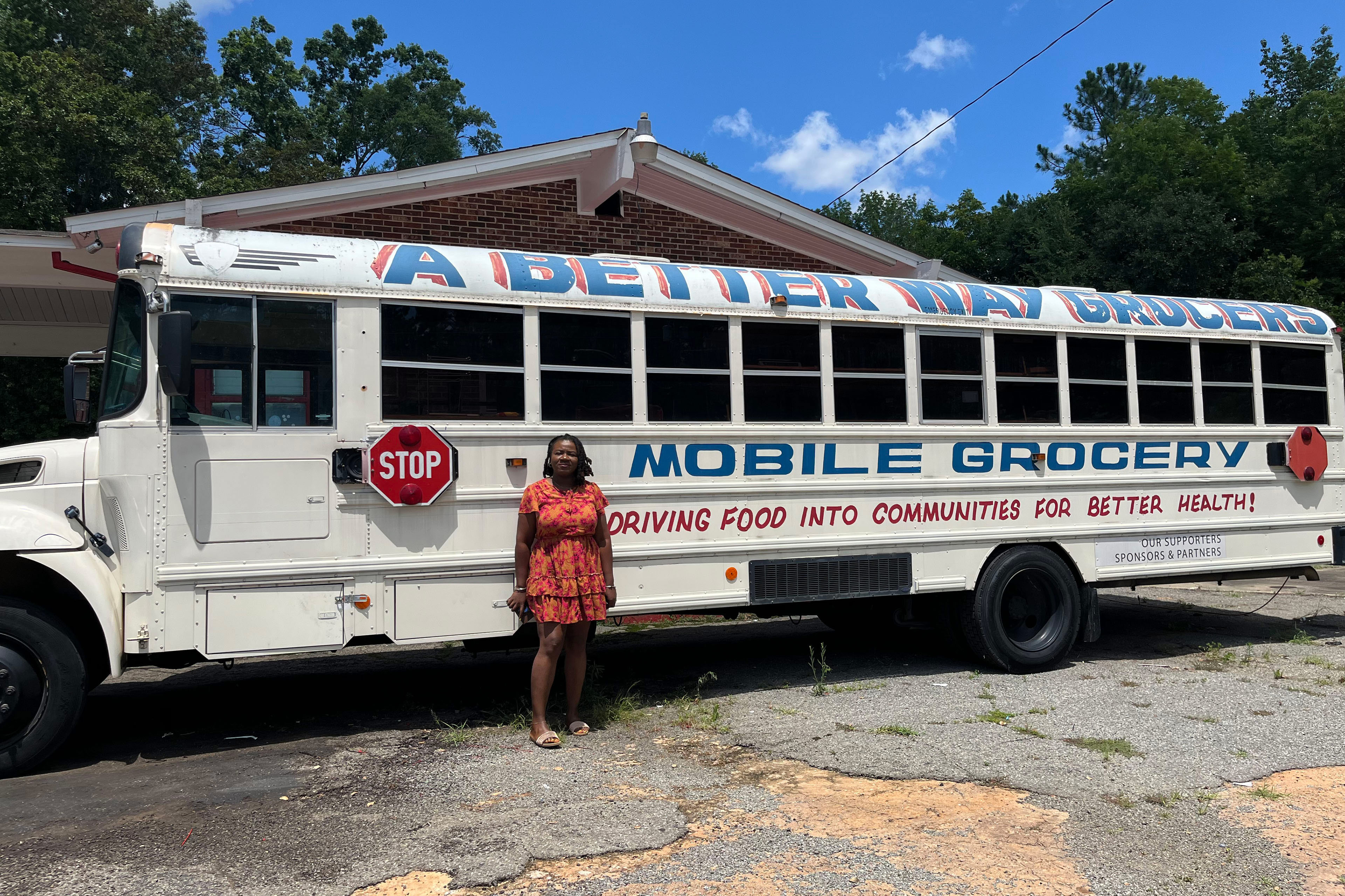 A photo of Terrell standing in front of a parked school bus painted white. There is writing on it that reads, "A Better Way Grocers," and "Mobile Grocery."