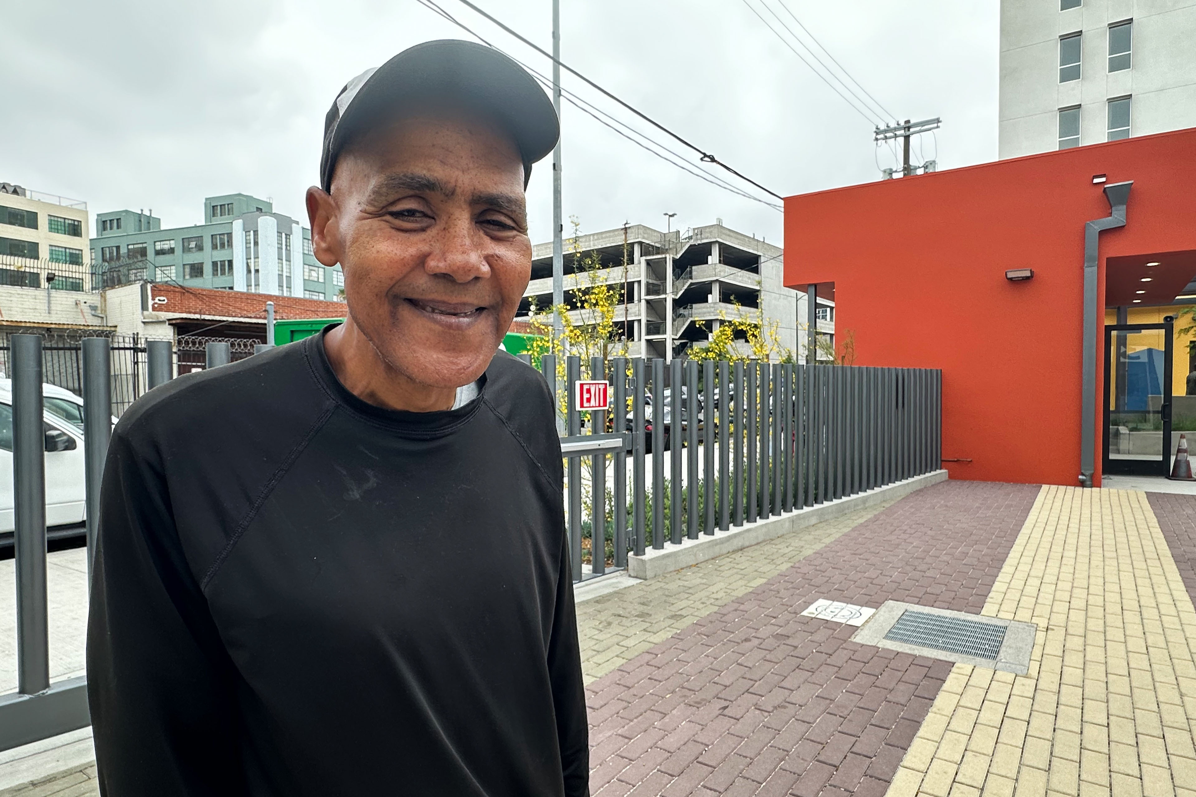 A portrait of a man wearing a baseball cap and a black shirt standing beside the Skid Row Care Campus building.