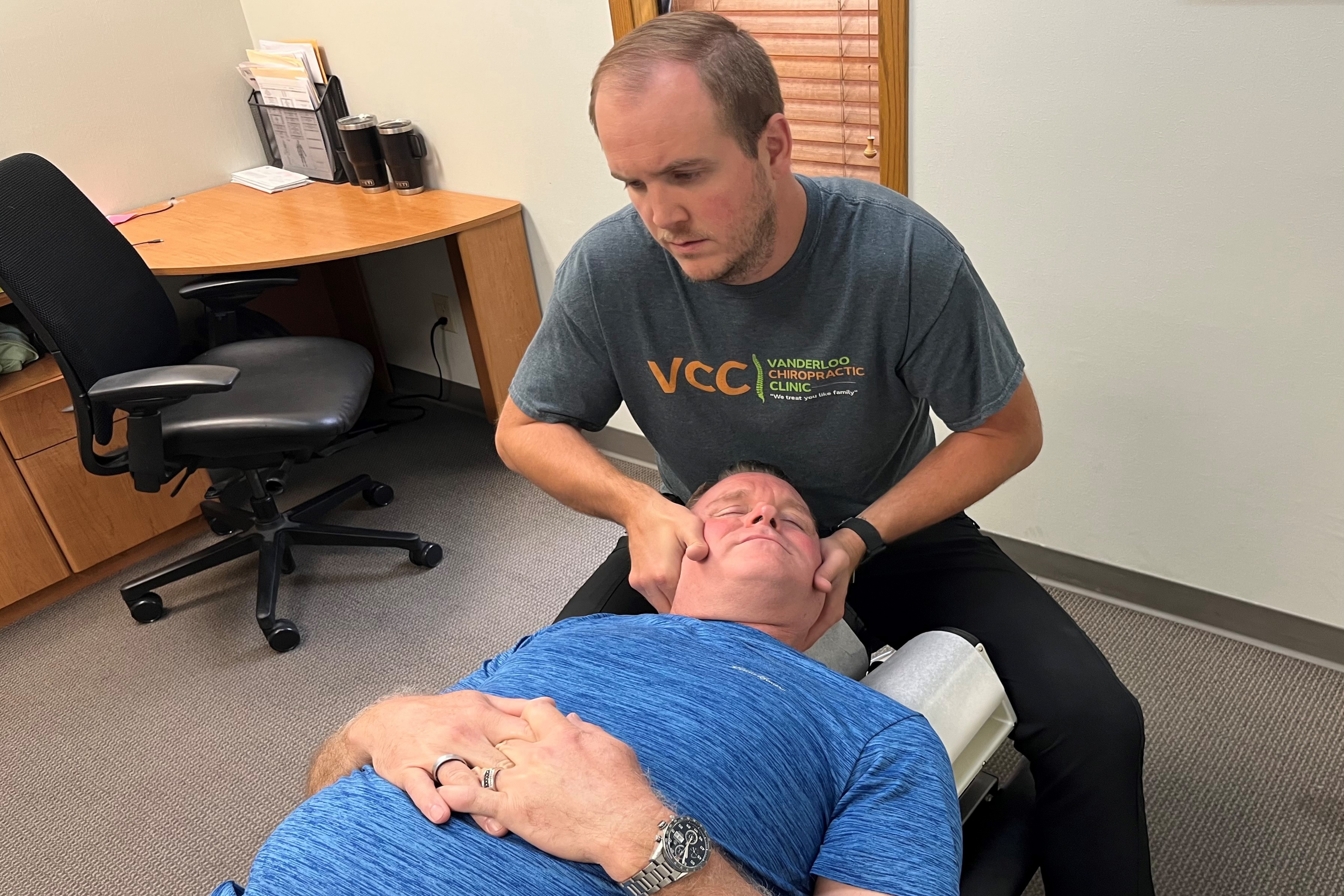 A man in a blue tshirt lays on a chiropractor bed while a man seated near his head cups either side of his face, bending his neck