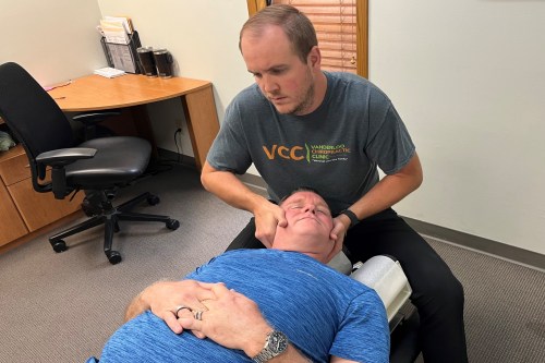 A man in a blue tshirt lays on a chiropractor bed while a man seated near his head cups either side of his face, bending his neck