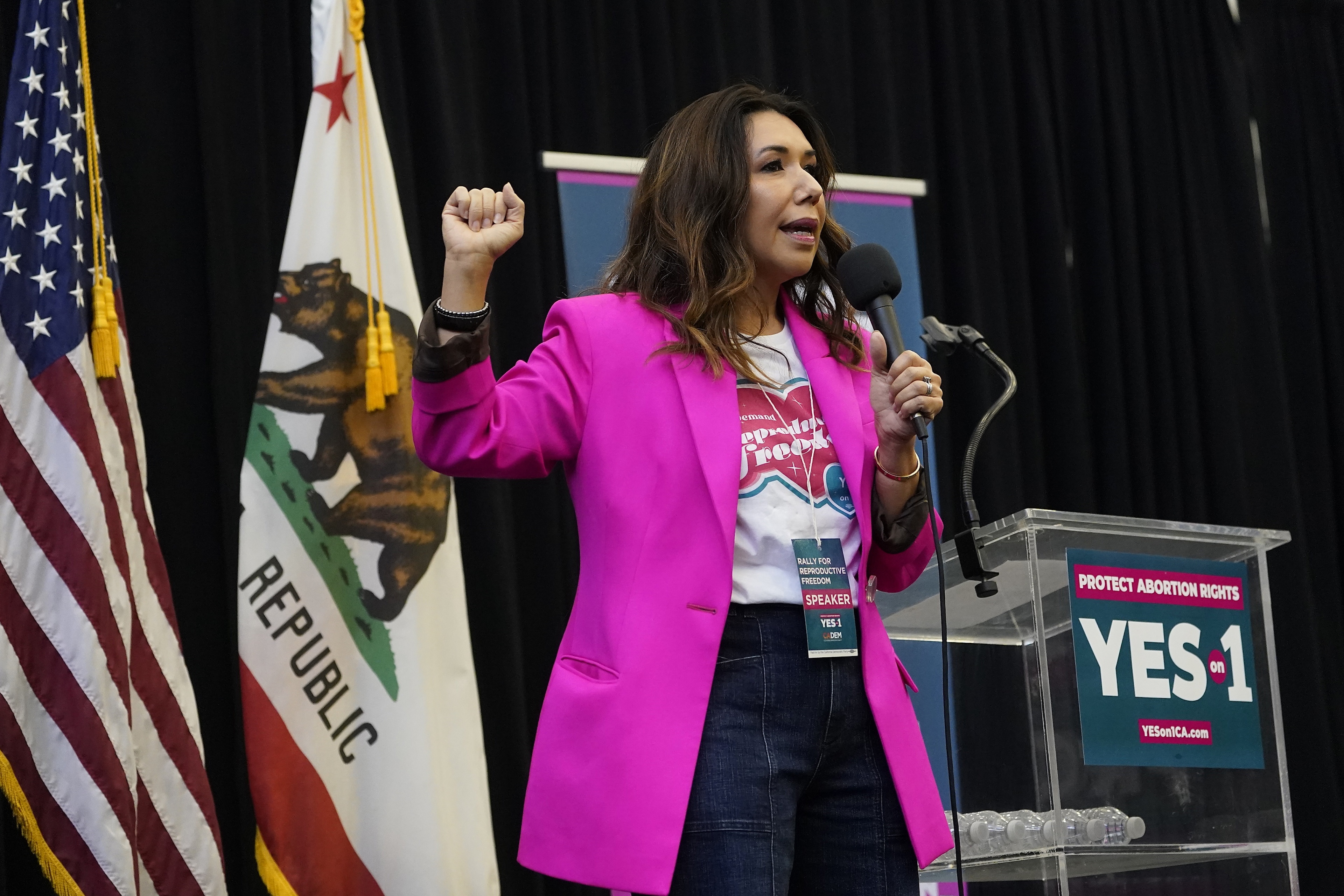 A woman in a bright pink blazer with a raised fist standing in front of a California and USA flag