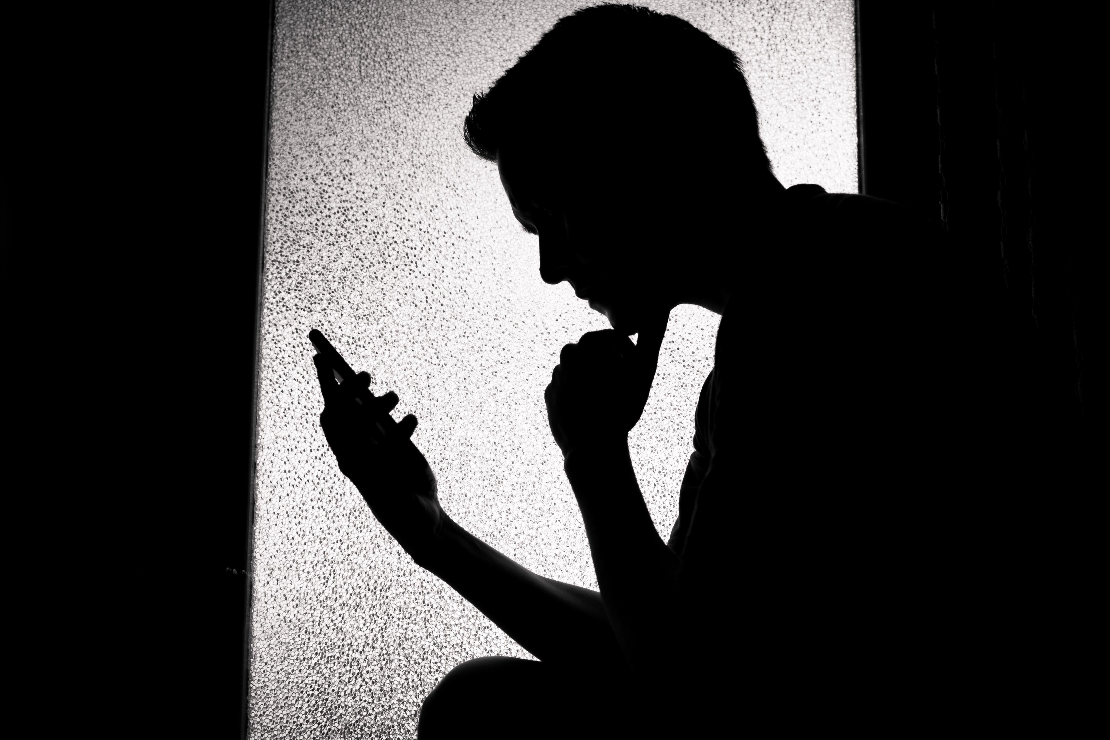 A photo of a young man in silhouette. He is looking at his phone.