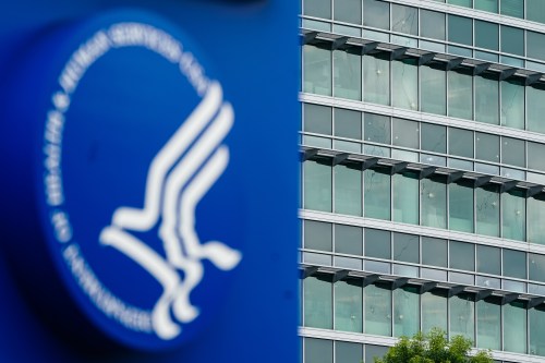A photo of a large office building. Its windows are riddled with bullet holes. In the foreground is a bright blue sign with an HHS logo.
