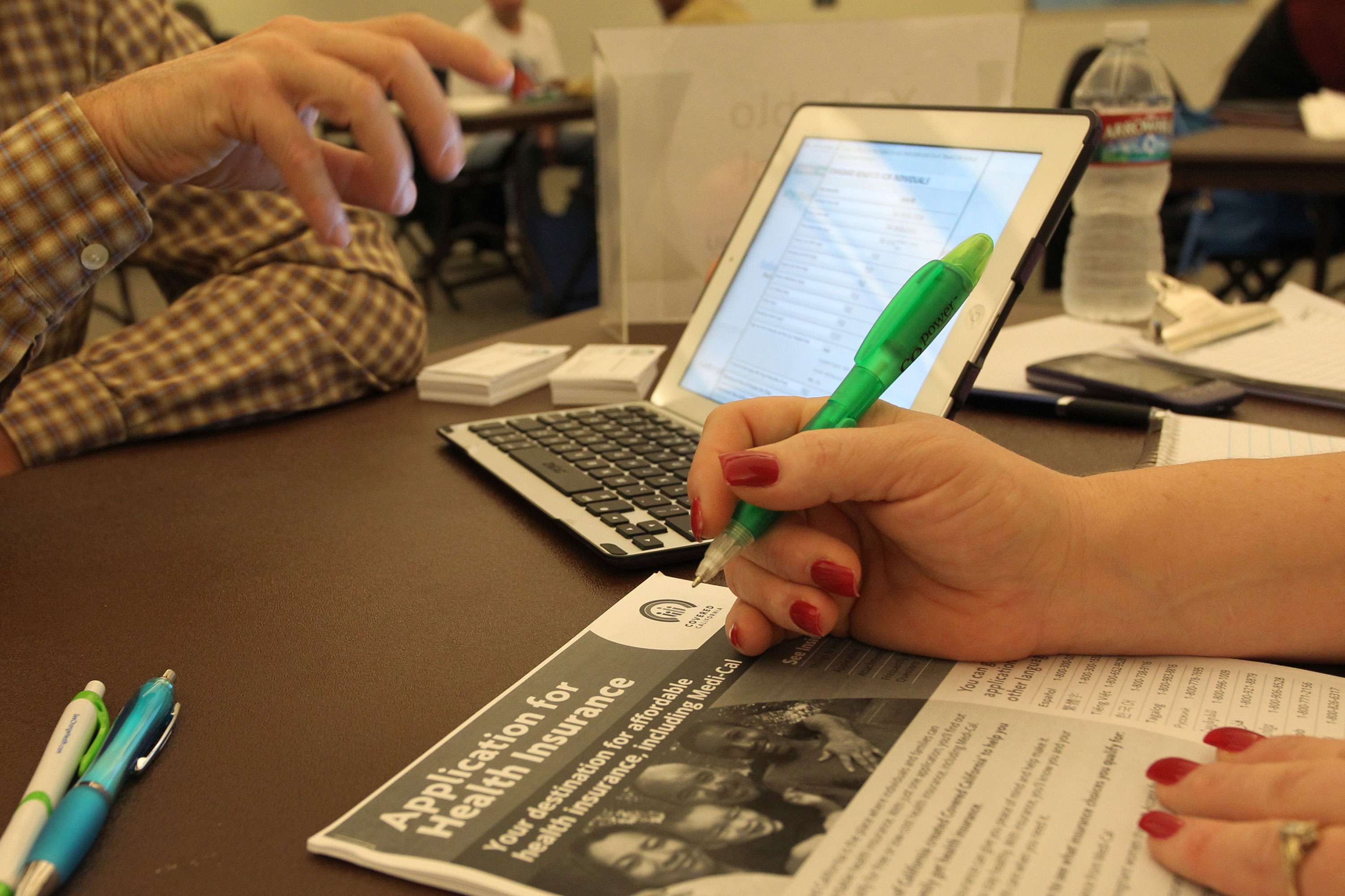 A photo of a laptop on a table. In the foreground is a woman holding a pen over a Covered California information packet.