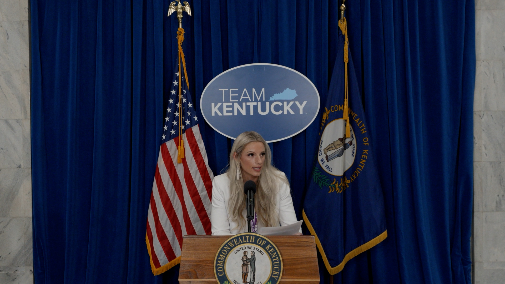 A photo of a woman speaking at a podium. A sign that reads "Team Kentucky" is behind her, as are the American and Kentucky flags.