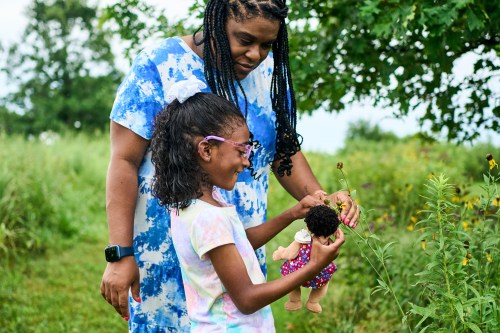 A photo of a mother with her daughter outside. The young girl is holding a doll and looking at flowers with her mom.