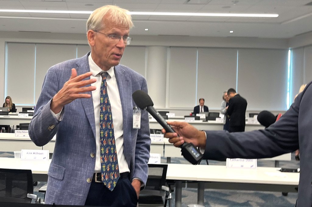 A man wearing a tie and blazer speaks into a microphone held by a reporter.