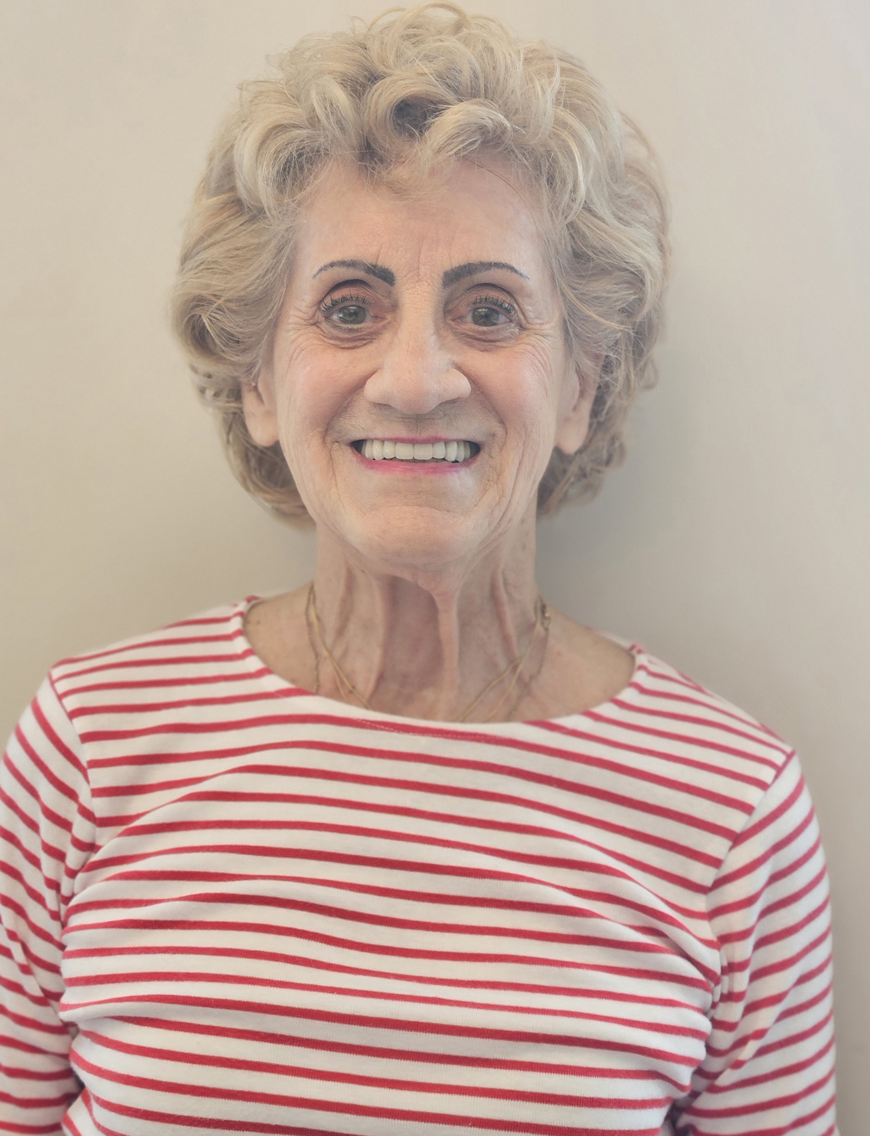 A portrait of a senior woman with short, curly white hair wearing a red-striped shirt and smiling broadly.