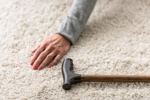 A cropped view of a senior woman's hand on the floor beside a cane on a carpeted floor.