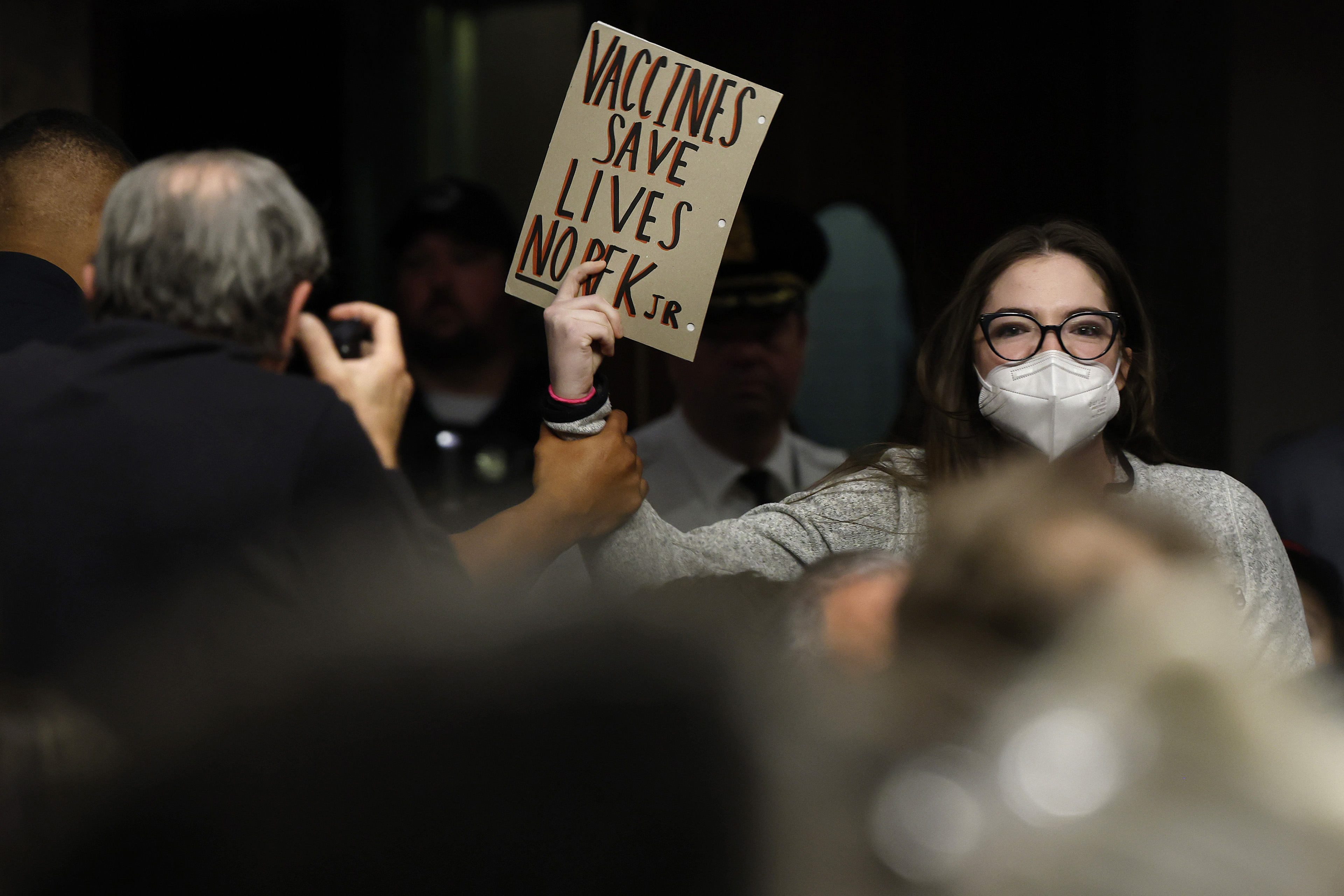 A woman wearing a mask and glasses holds a protest sign as she is led out of a hearing at the Dirksen Senate Office Building