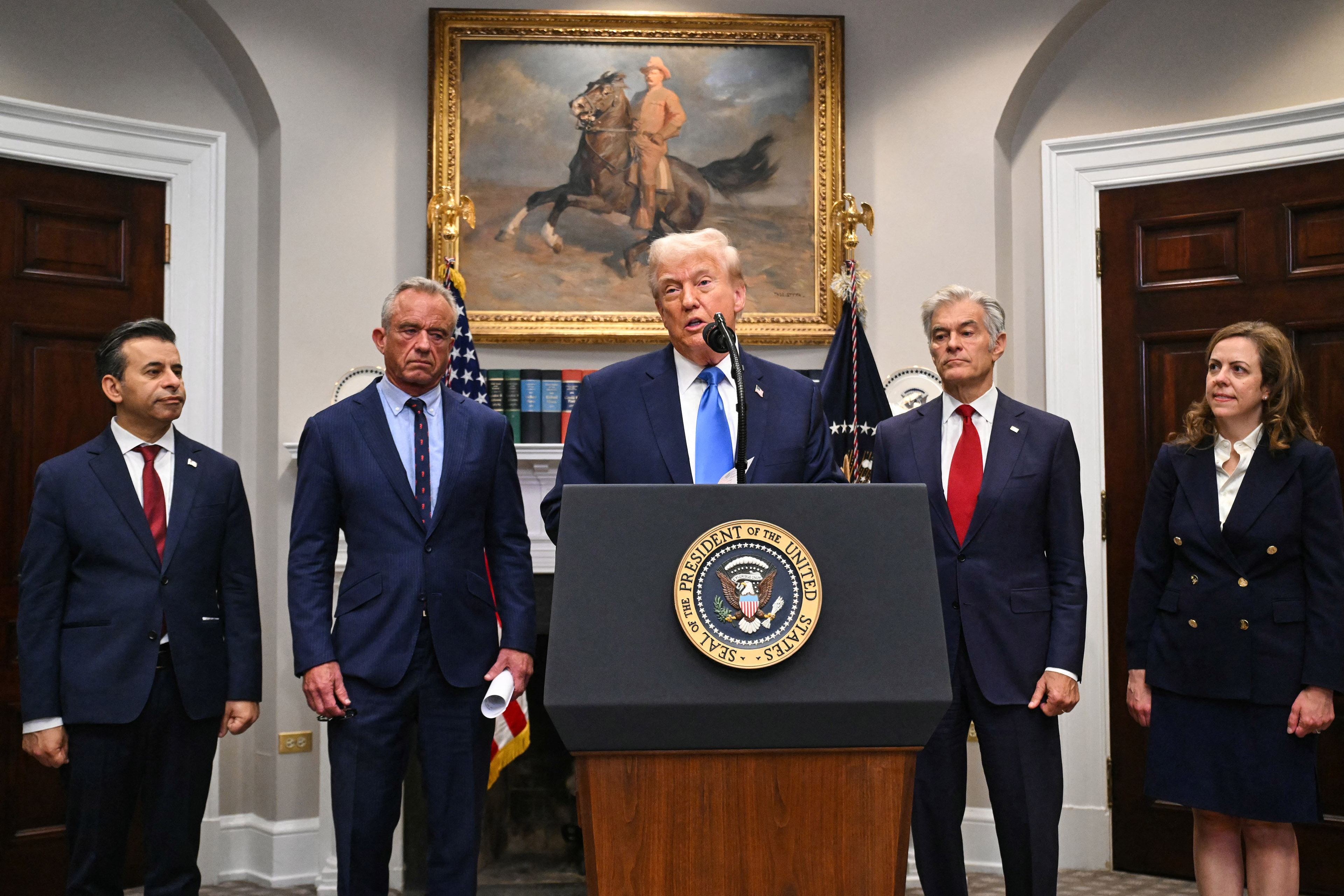 A photo of President Donald standing at a podium speaking into a microphone flanked by four other people