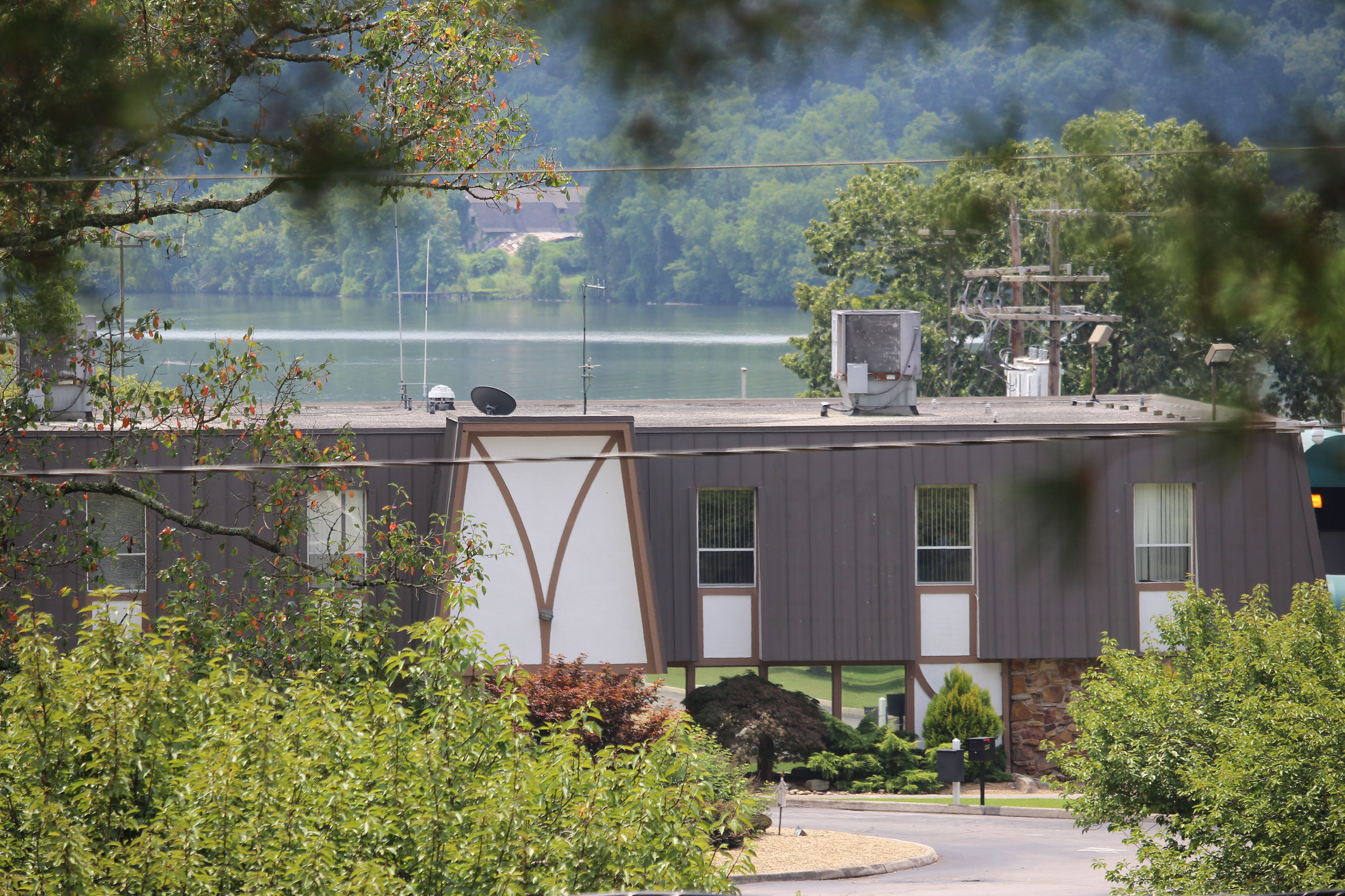 A low building is shown surrounded by trees and with a river behind it.