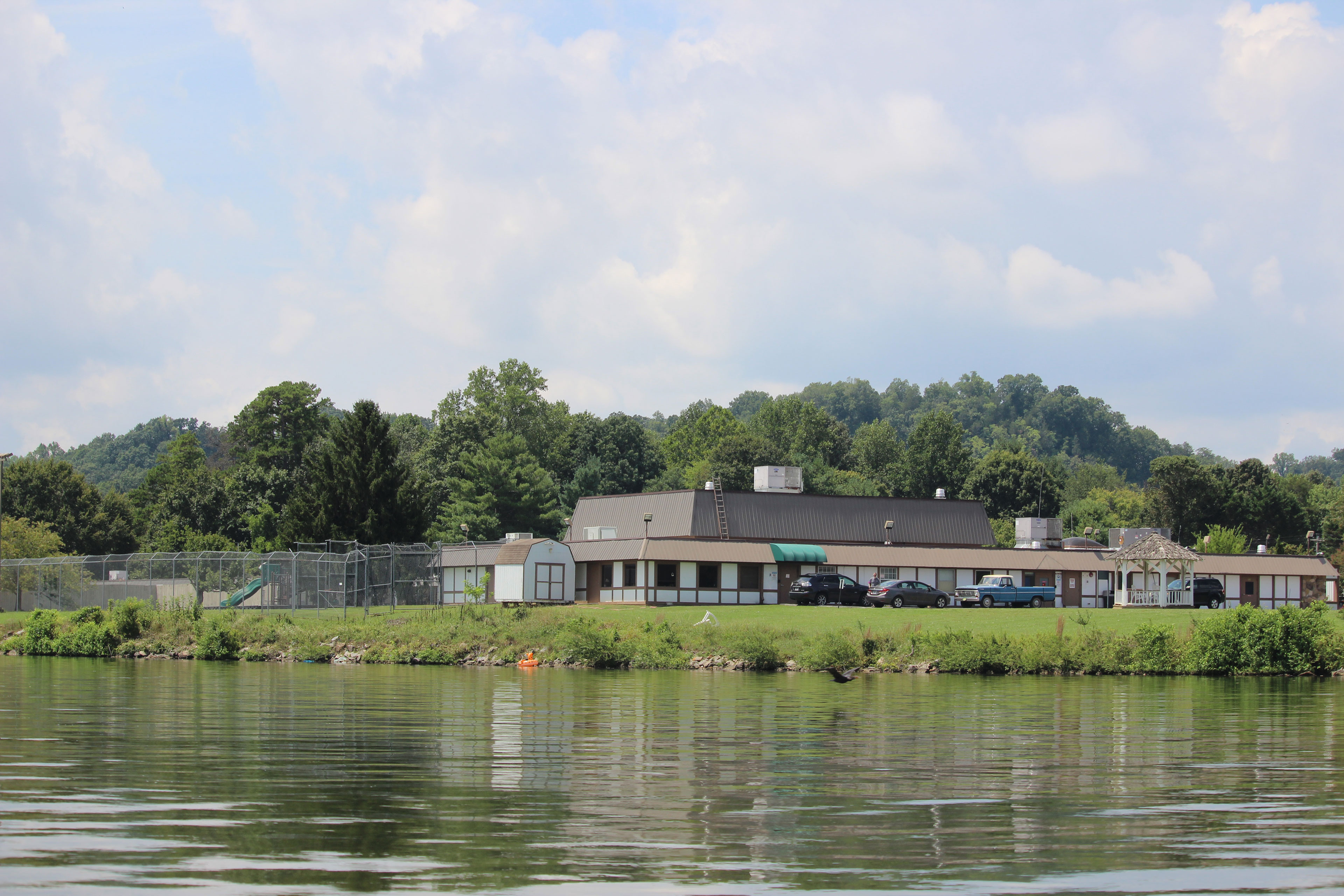 In a photo taken from a craft on a river, a single-story building building sits near the water. There is a fenced-in playground, a shed, a gazebo, and a small parking lot surrounding the building. Trees can be seen in the distance behind the building and its grounds.