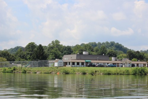 In a photo taken from a craft on a river, a single-story building building sits near the water. There is a fenced-in playground, a shed, a gazebo, and a small parking lot surrounding the building. Trees can be seen in the distance behind the building and its grounds.