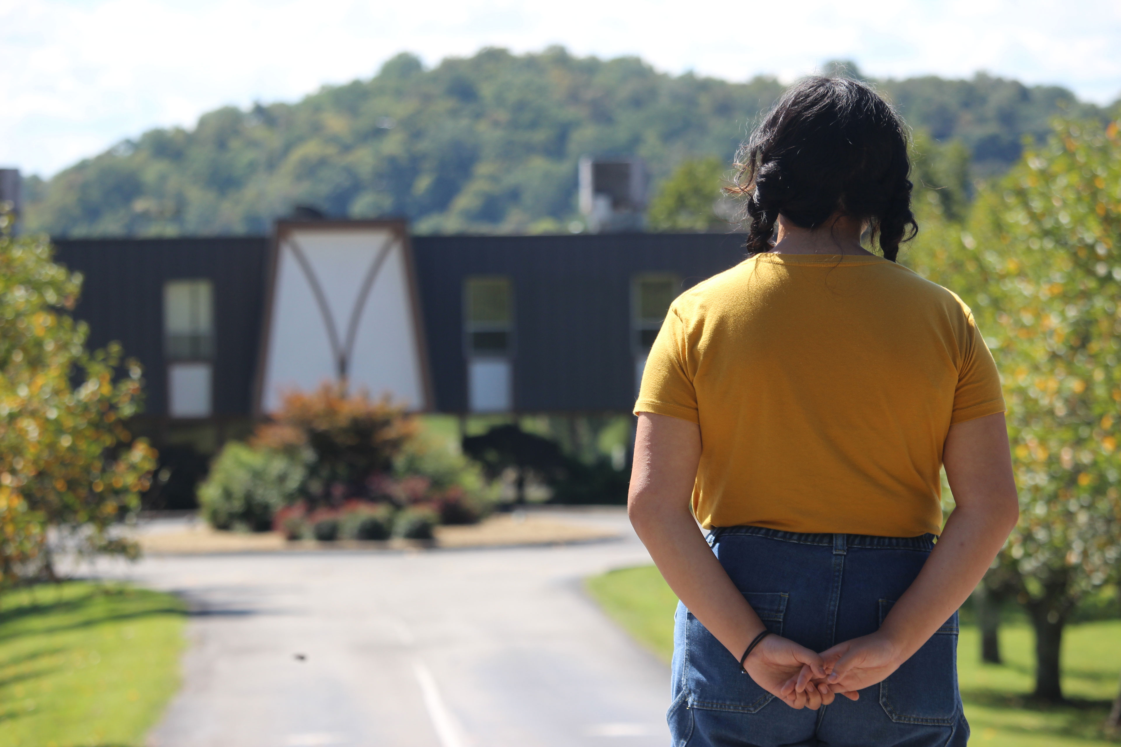 A woman wearing a yellow t-shirt and jeans stands with her back to the camera and looks an a building at the other end of a long driveway. Her dark hair is in two braids and her hands are clasped behind her back.