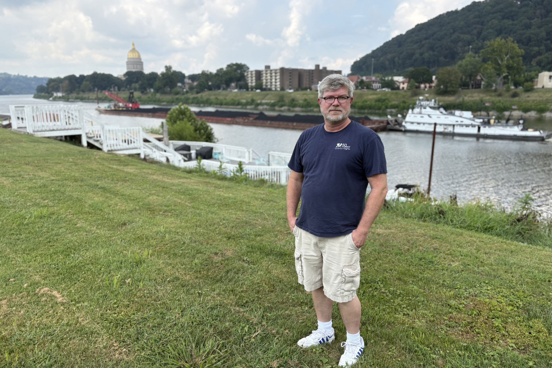 A man with sandy-gray hair and beard, wearing glasses, a navy blue t-shirt, khaki shorts, and sneakers stands on grass and looks at the camera. His hands are in his pockets. Behind him is a river, a boat on the river, and buildings on the opposite shore, including a golden dome in the distance.