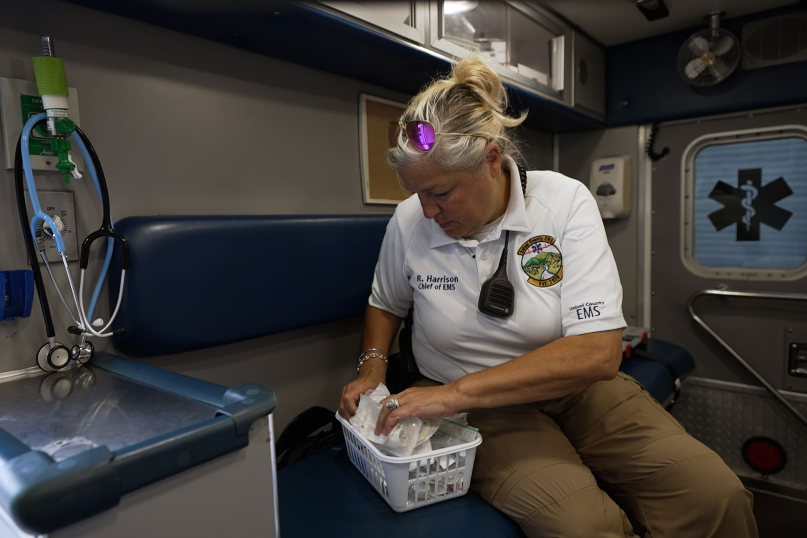 A woman with her blonde hair up in ponytail organizes items in a plastic container while sitting inside an ambulance. Her white polo shirt reads "R. Harrison, Chief of EMS" and has an emblem on the front. The left sleeve reads "Unicoi County EMS".