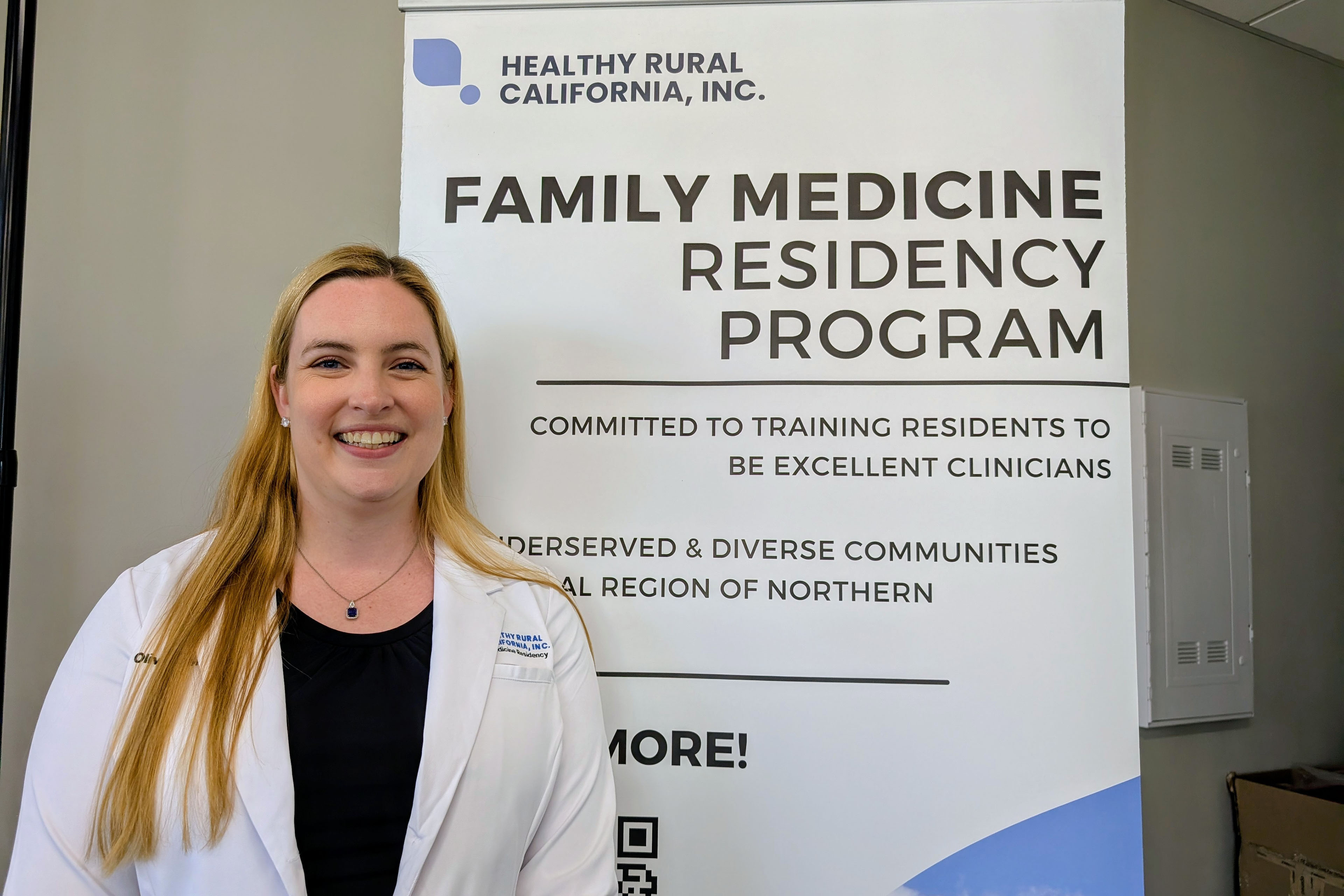 A photo of a female doctor wearing a white coat standing beside a sign that says: "Healthy Rural California, Inc. / Family medicine residency program / committed to training residents to be excellent clinicians."