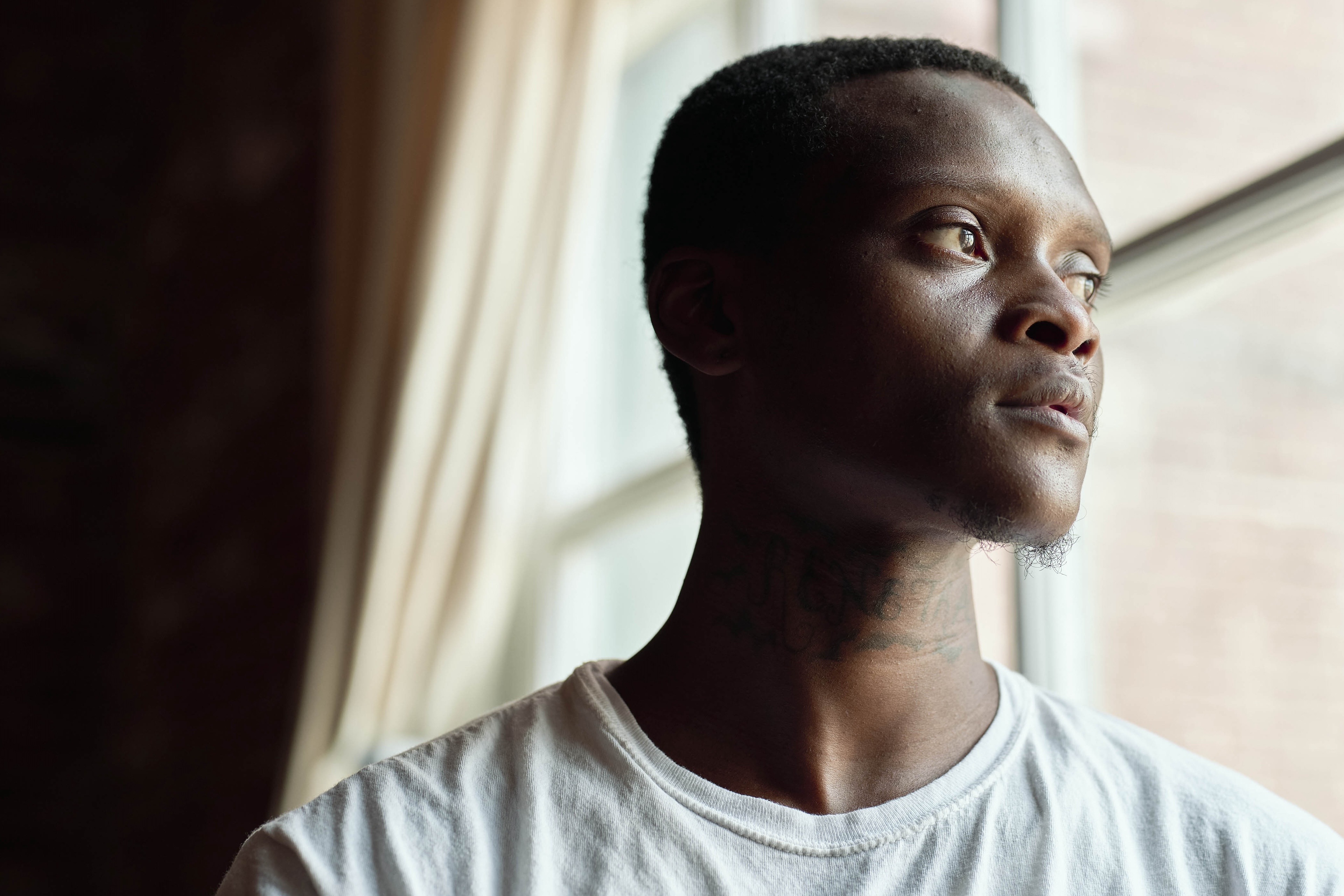 A man in a white tshirt stands in front of a window looking outwards