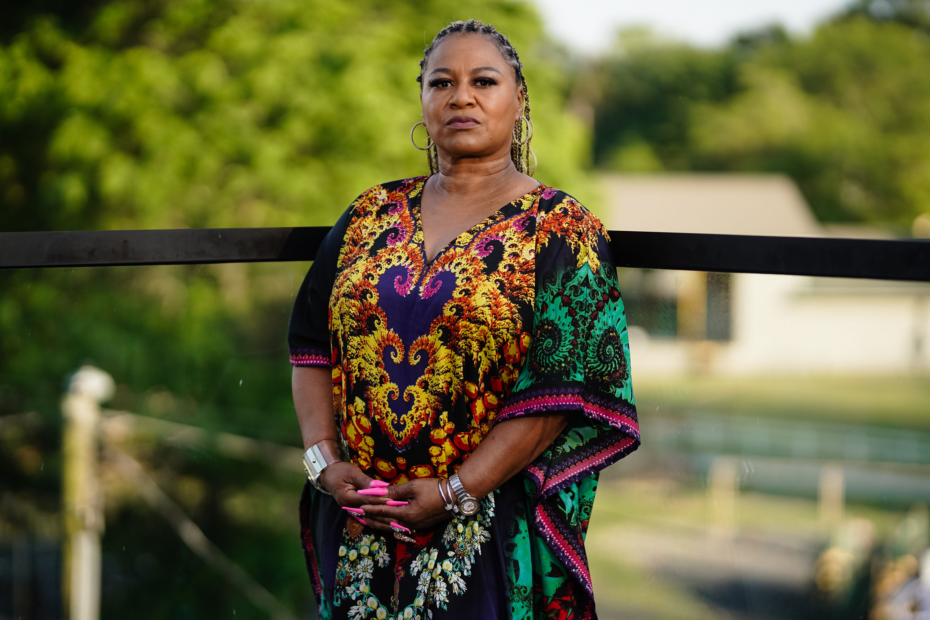 A portrait of a woman looking directly, confidently, straight at the camera. She stands with her hands held together in front of her, outside.