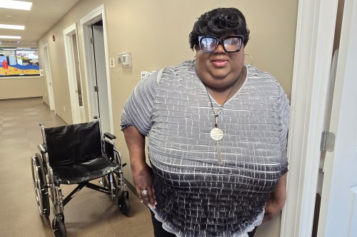 A photo of a woman standing for a portrait in a hallway. A wheelchair is behind her.