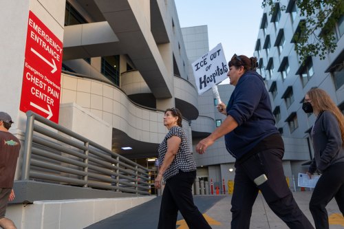 Four people are in frame walking past a sign with white lettering on a red background reading "Emergency Entrance" and "Emergency Department Chest Pain Center" each with arrows pointing right. One person carries a handwritten sign that reads "Keep ICE Out of Hospitals."