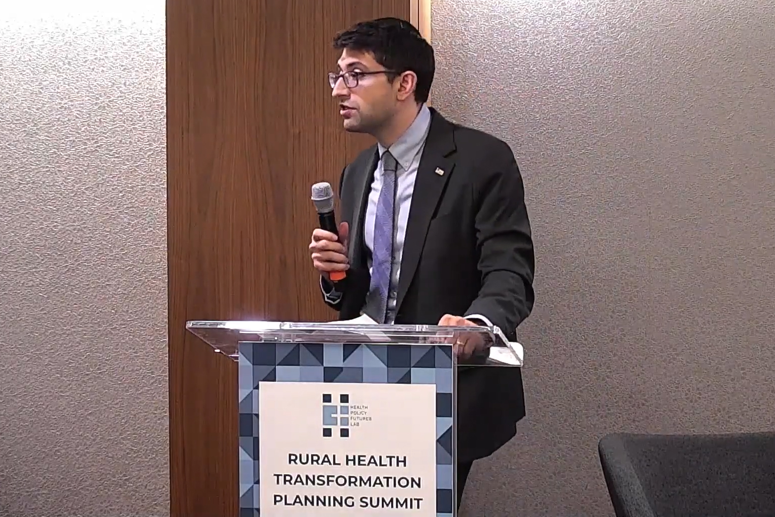 A photo of a man standing at a podium and speaking into a microphone.