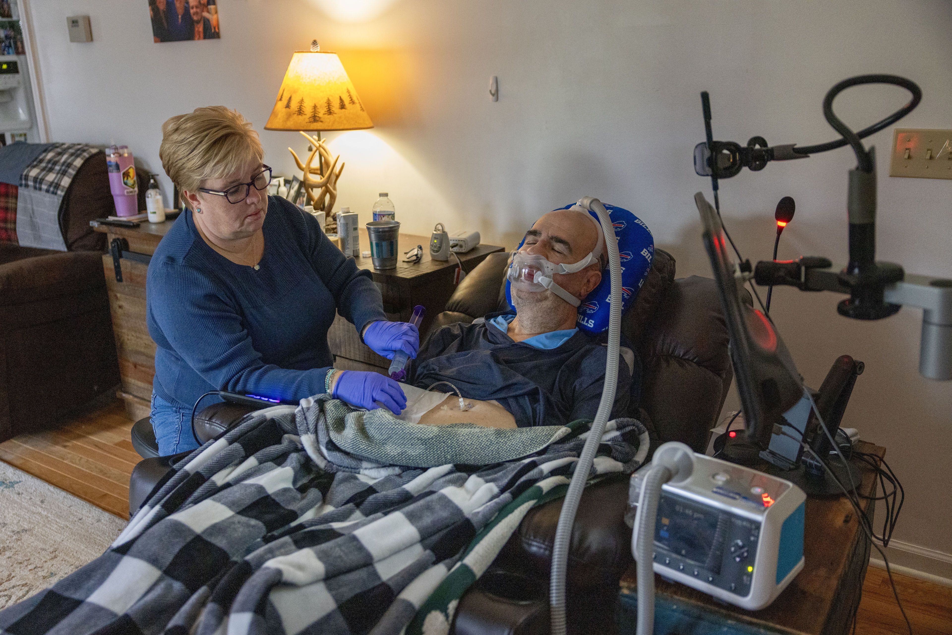 A woman wearing blue nitrile gloves helps a man who is sitting in a recliner next to her wearing a ventilator