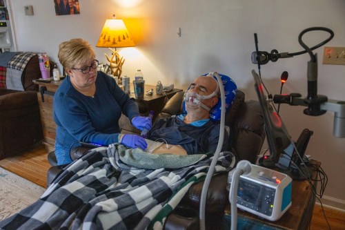 A woman wearing blue nitrile gloves helps a man who is sitting in a recliner next to her wearing a ventilator