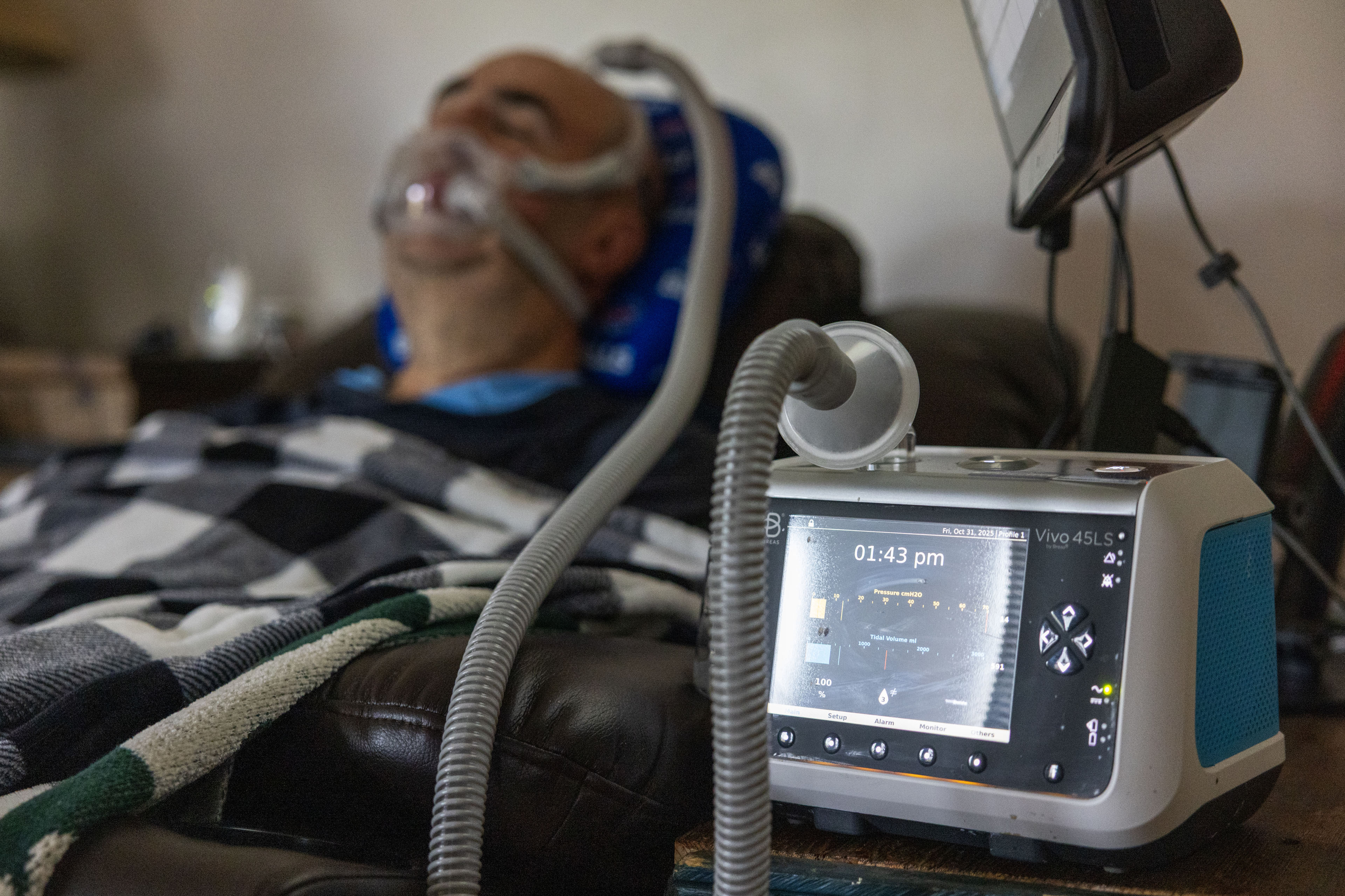 A ventilator machine is in the foreground, while a man wearing a ventilator mask is in the background