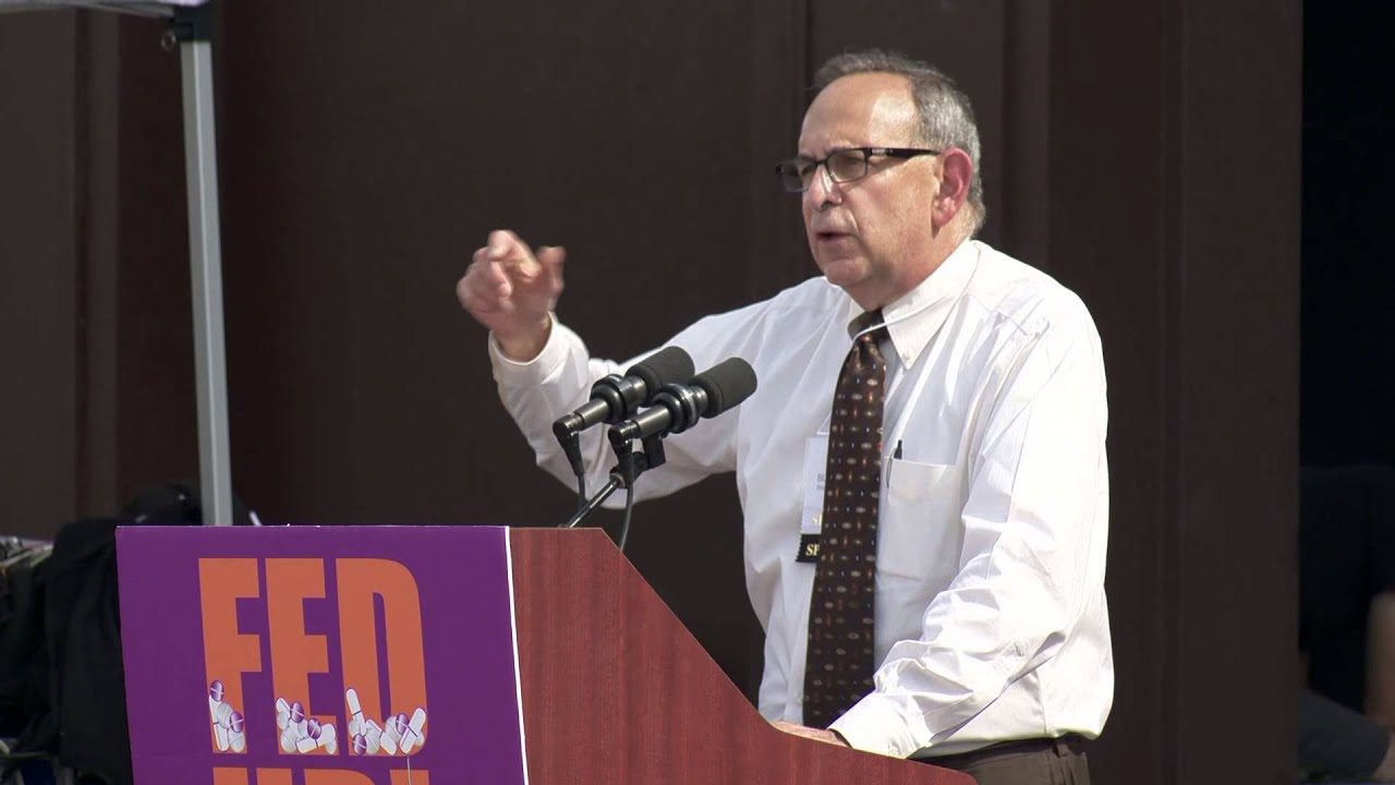 A man wearing a collared shirt and tie speaks from behind a lectern. 
