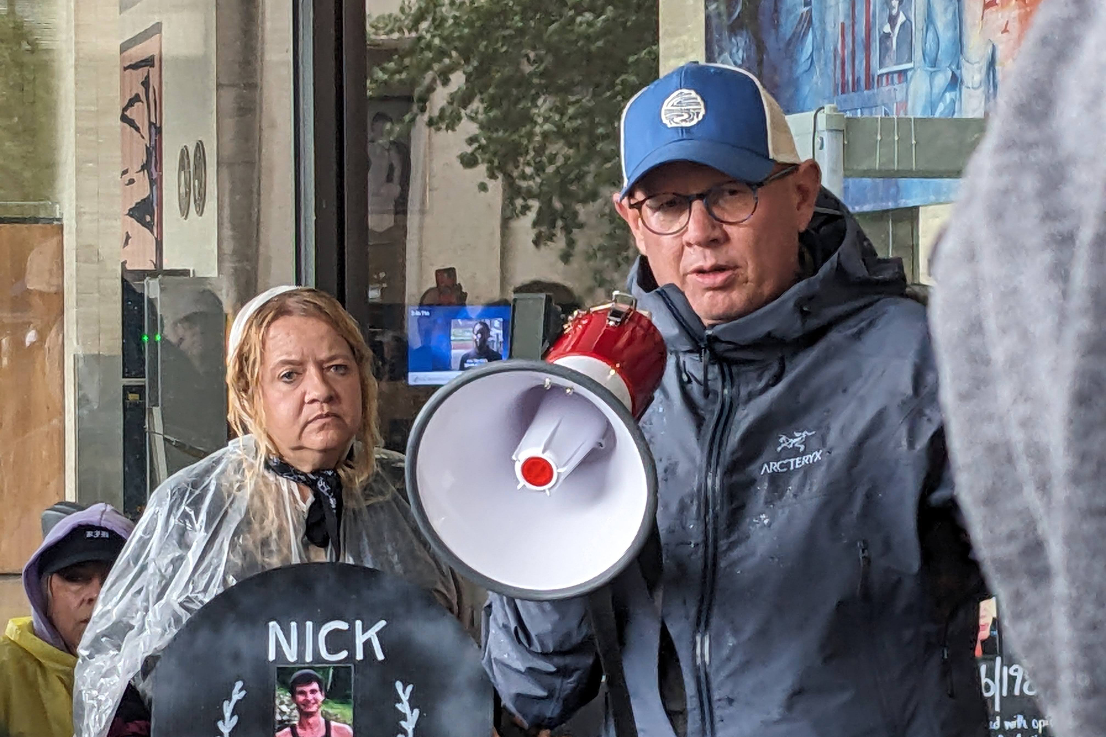A man in a raincoat and baseball cap speaks into a megaphone.