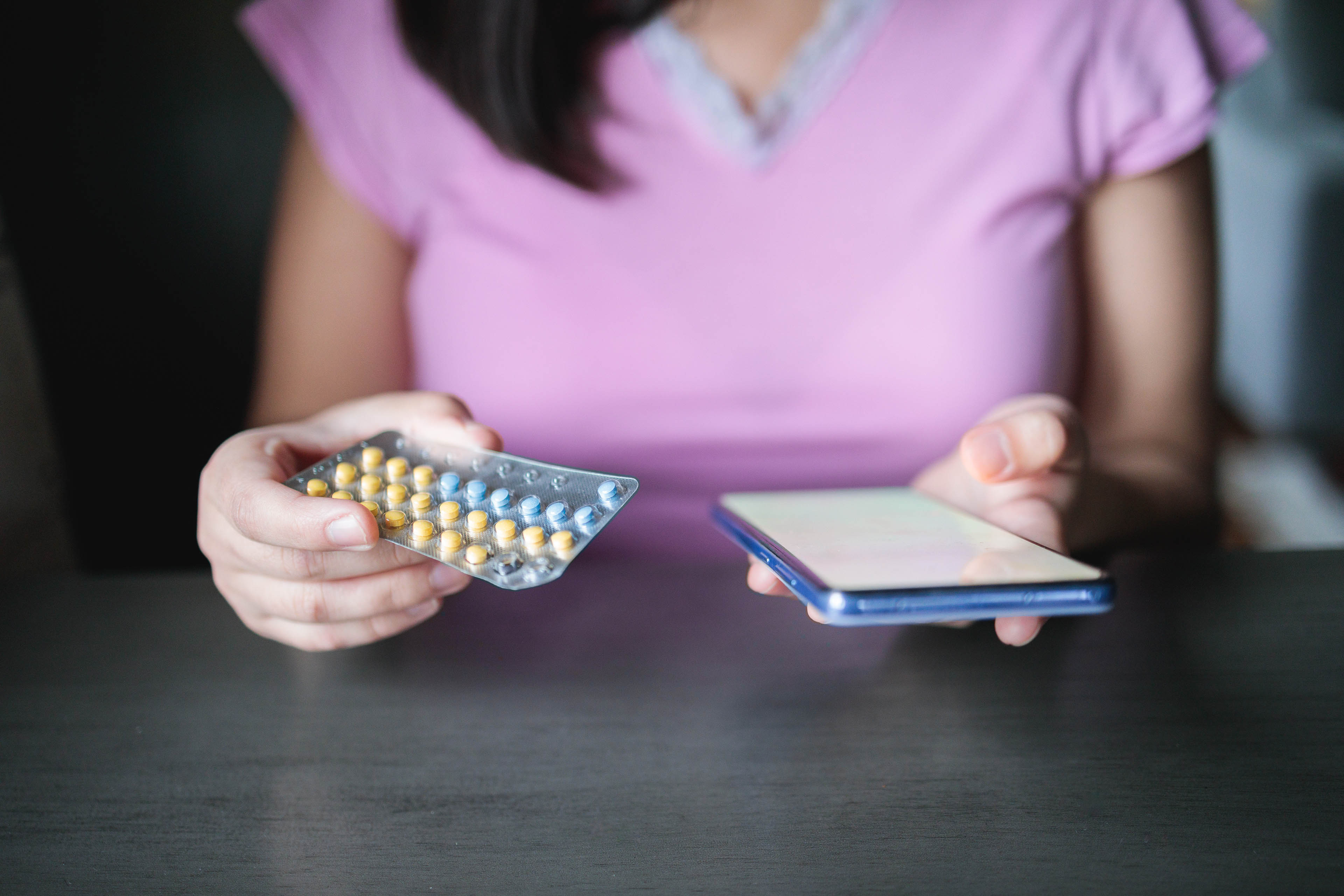 A photo from the shoulders down of a woman holding her a birth control pill pack in her left hand and a mobile phone in her right.