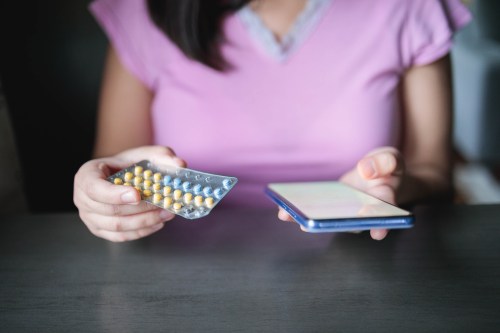 A photo from the shoulders down of a woman holding her a birth control pill pack in her left hand and a mobile phone in her right.