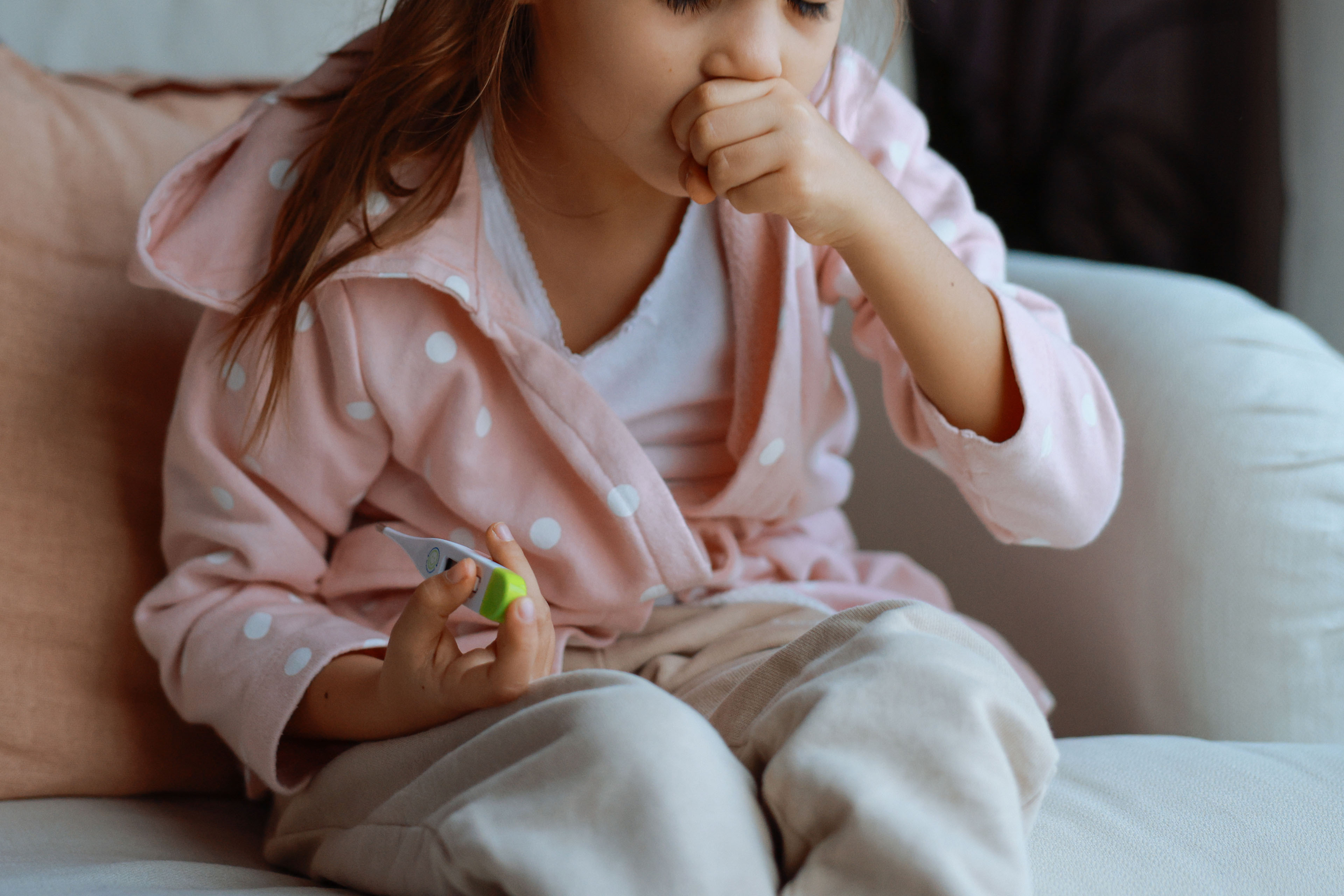 A closely cropped shot of a preschool-aged girl coughing while holding a thermometer.