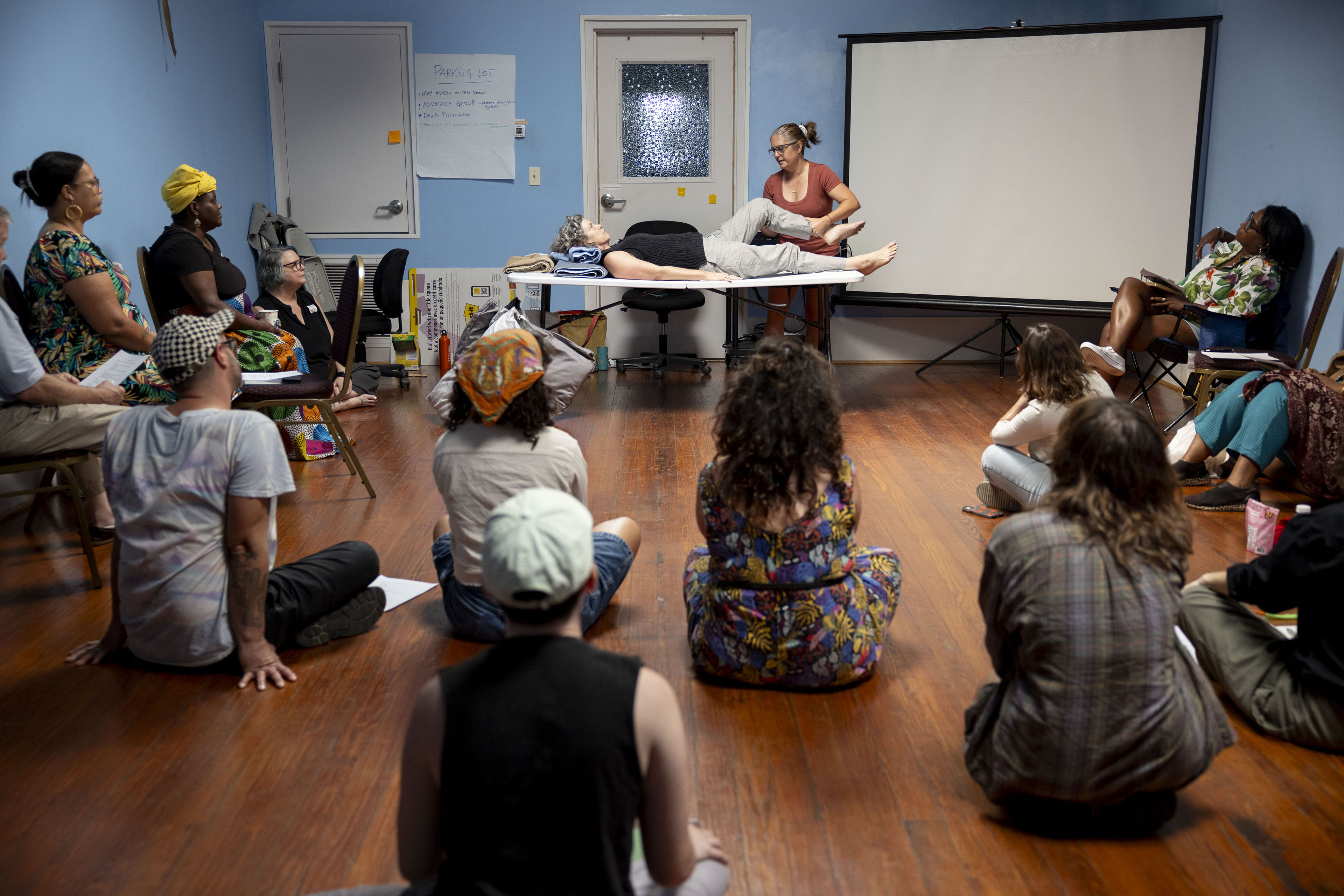 A group of people sit on the floor while they watch a presenter show home caregiving techniques.