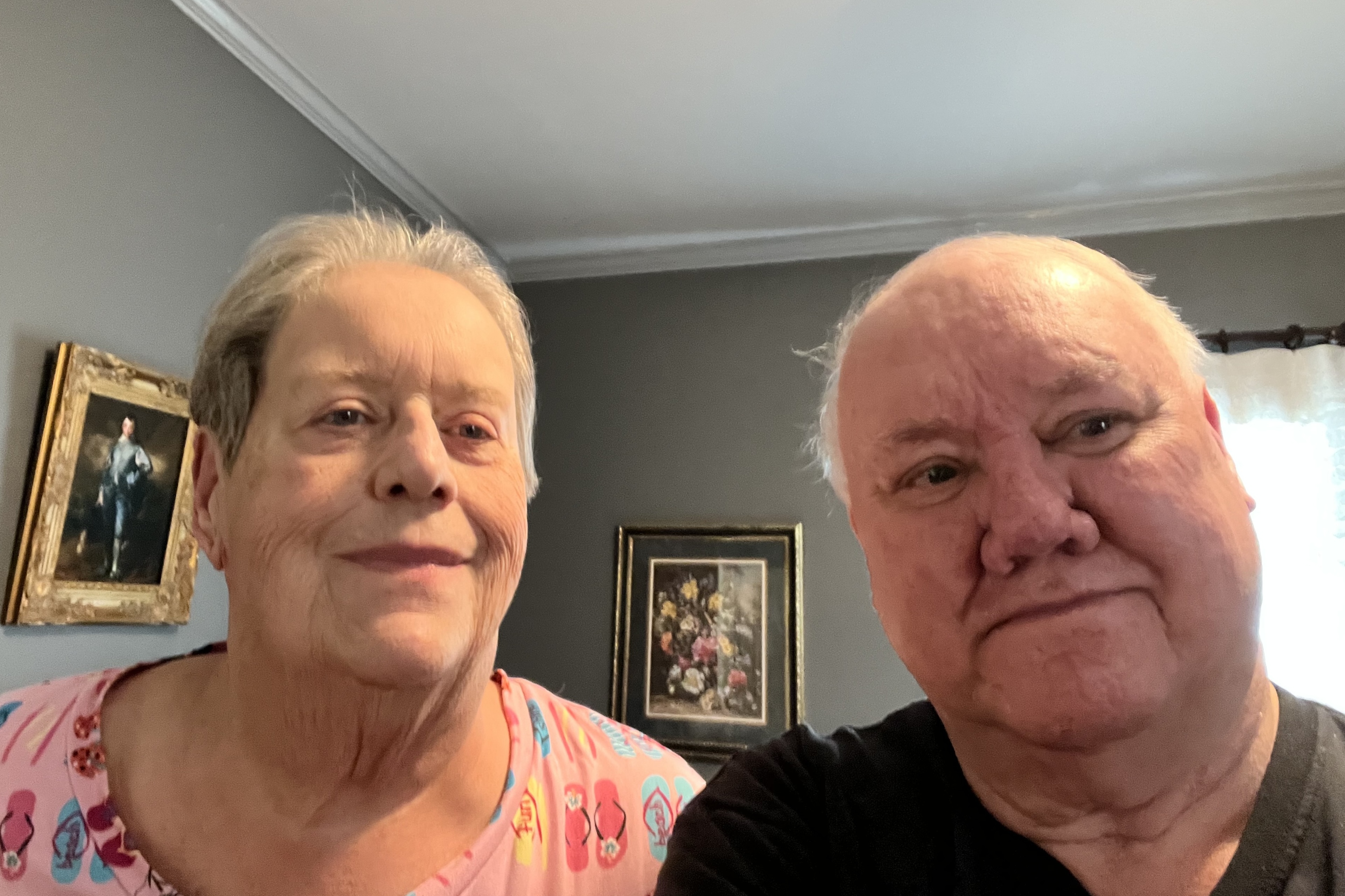 A senior man and woman seated in a room with several framed images behind them
