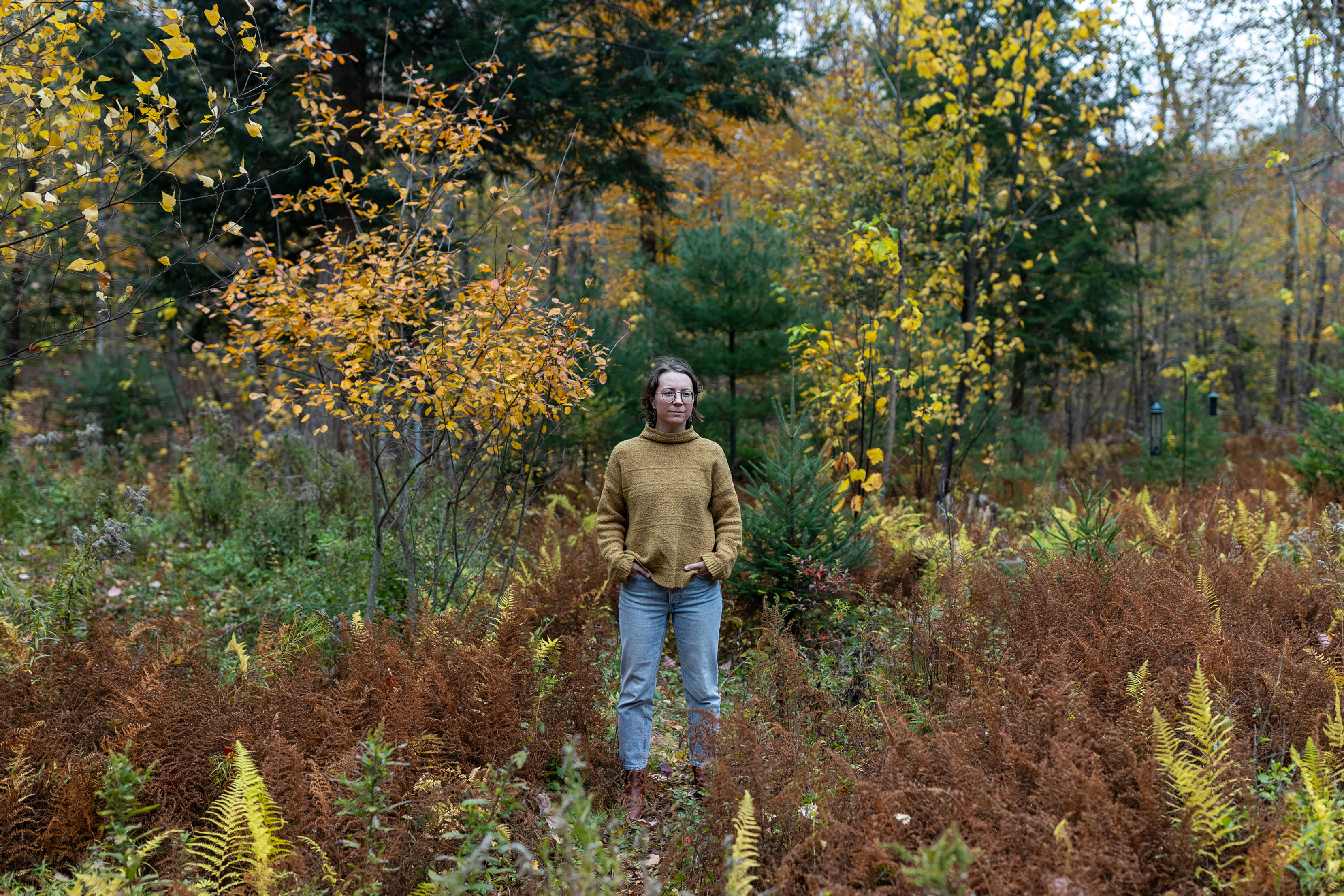 A photo of a woman standing in a meadow in autumn. The foliage around her is turning orange and yellow.