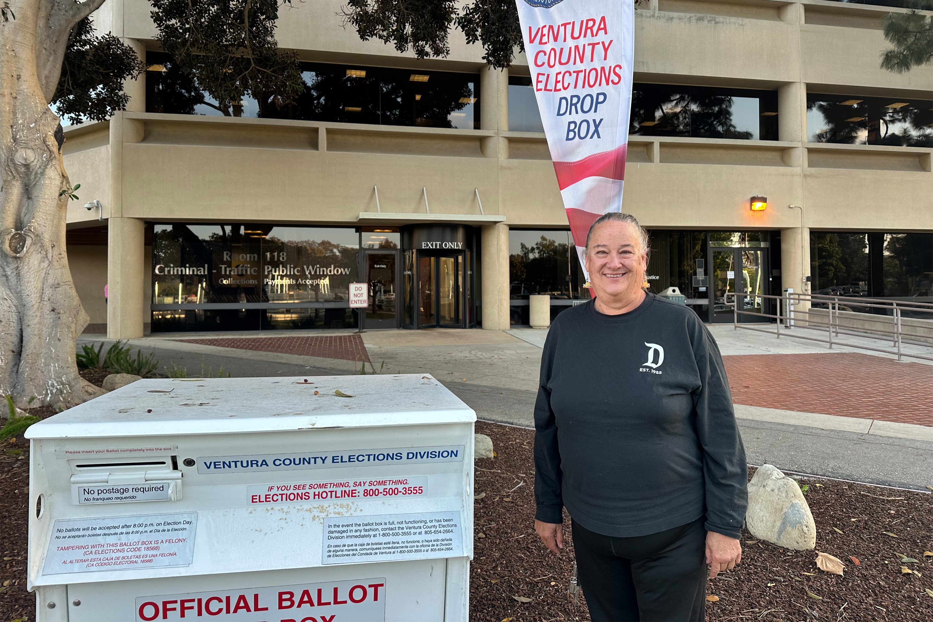A woman stands beside a ballot box, smiling.