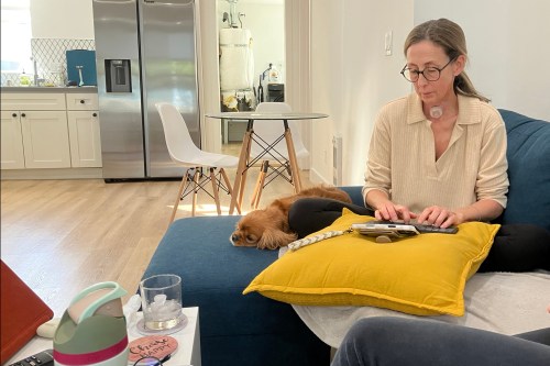 A woman sits on a blue couch typing on a keyboard before a phone. A small dog lays beside her.