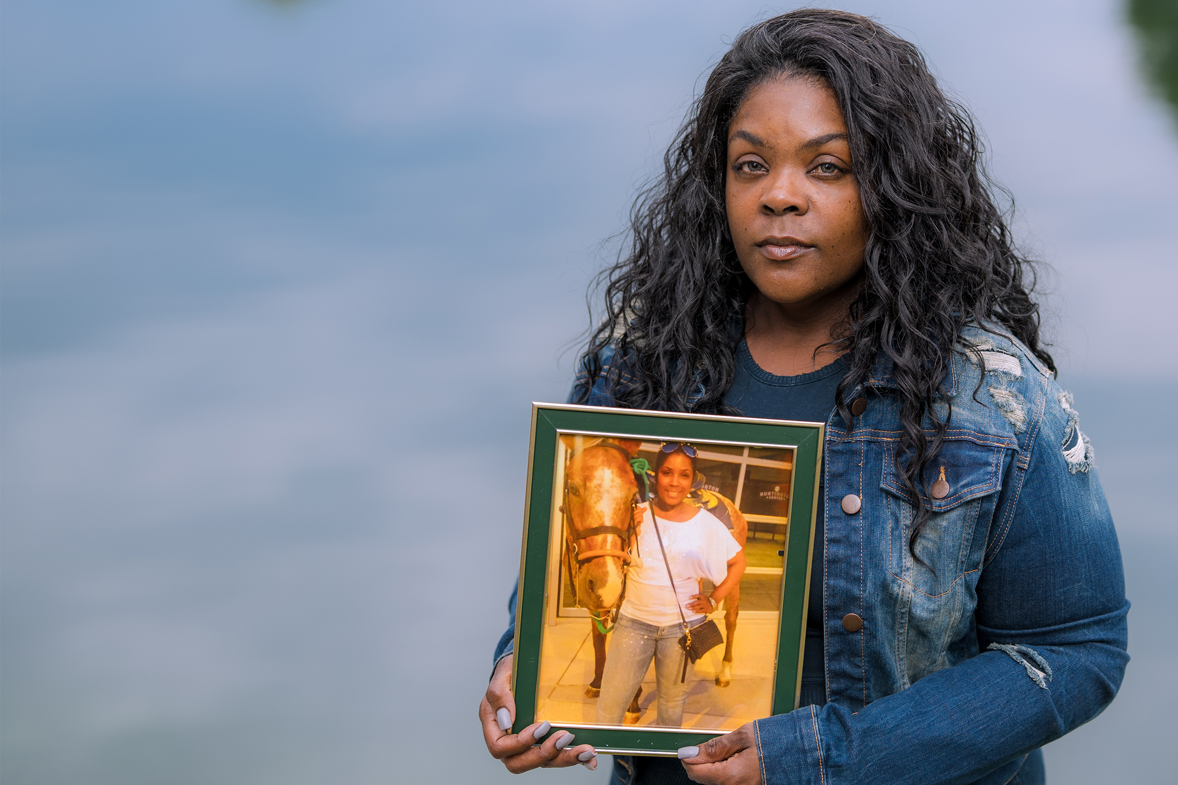 A photo of a Black woman standing by water. She holds a framed photo of her deceased mother.