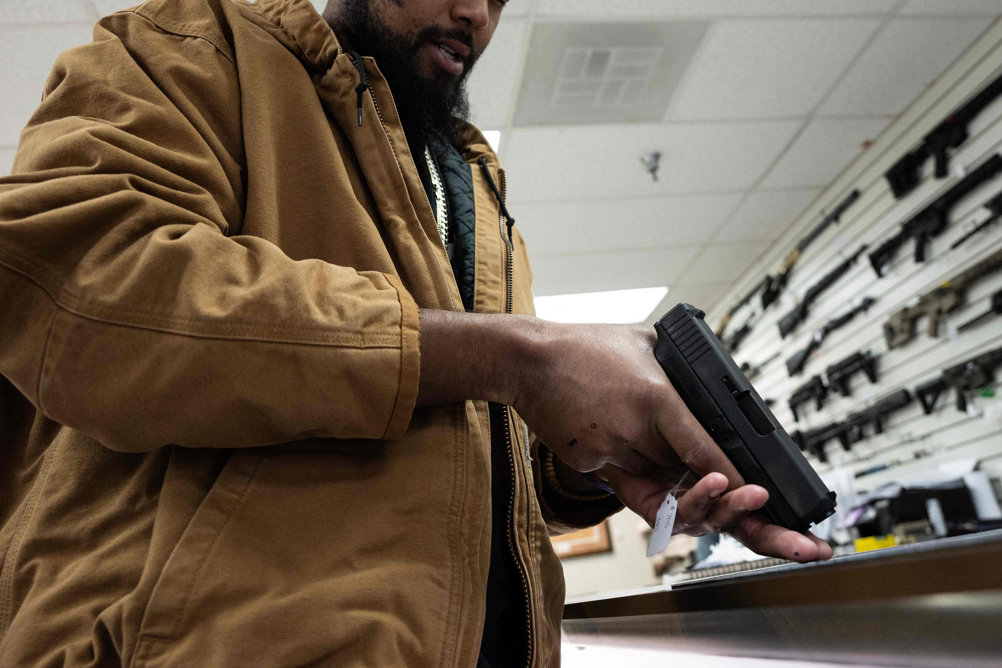 A photo of a man inspecting a pistol at a gun shop. Long guns are seen on the wall behind him.