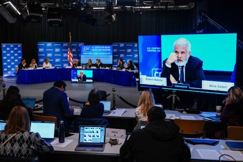 A photo of the ACIP hearing from the press section. Reporters type on laptops in the foreground. In front of them is a large TV screen showing Robert Malone speaking. The full panel is seen behind the screen.