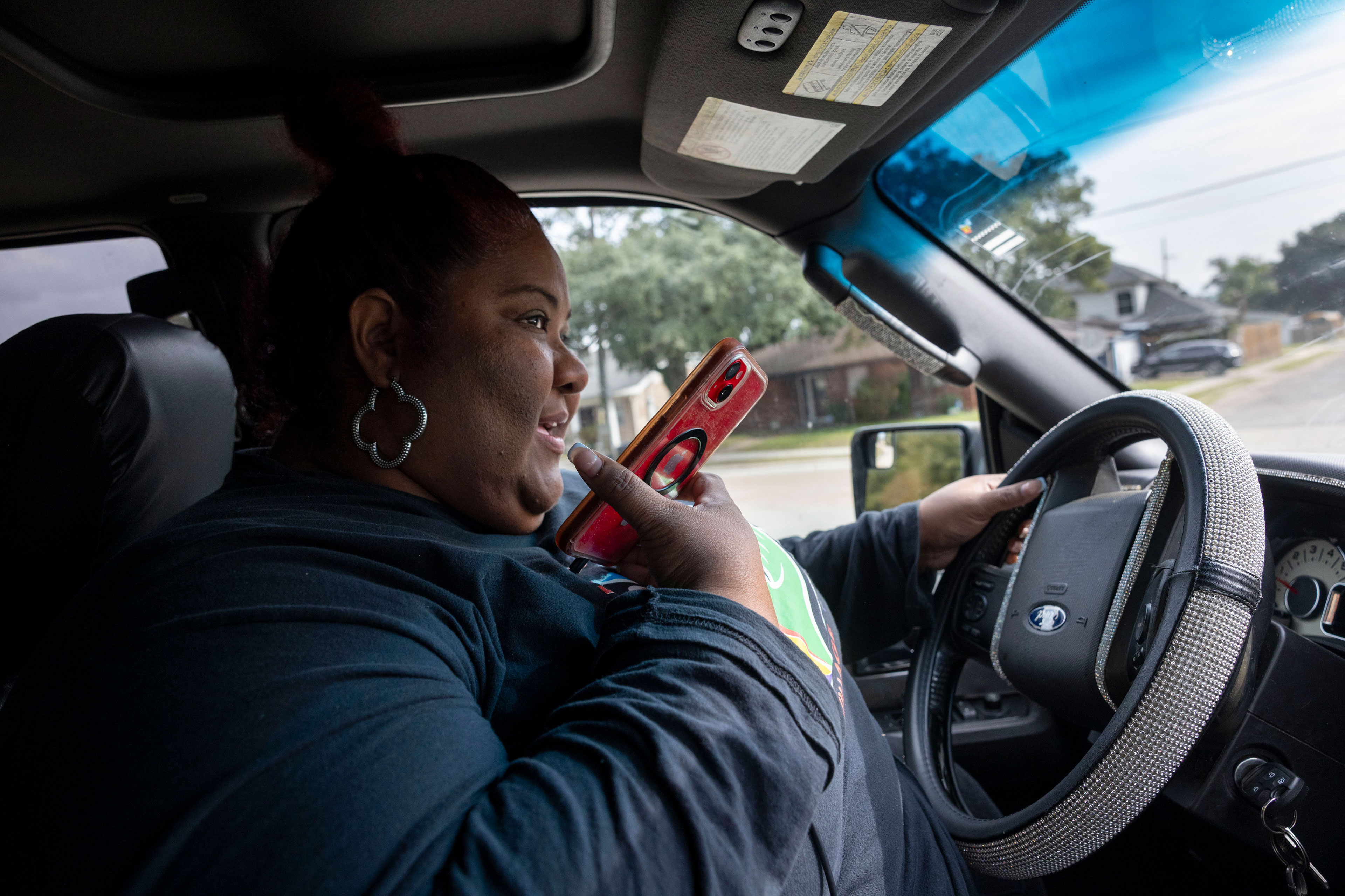 A woman in the driver's seat of a car holds the steering wheel in her left hand, and speaks into a cell phone held near her face in her right hand.