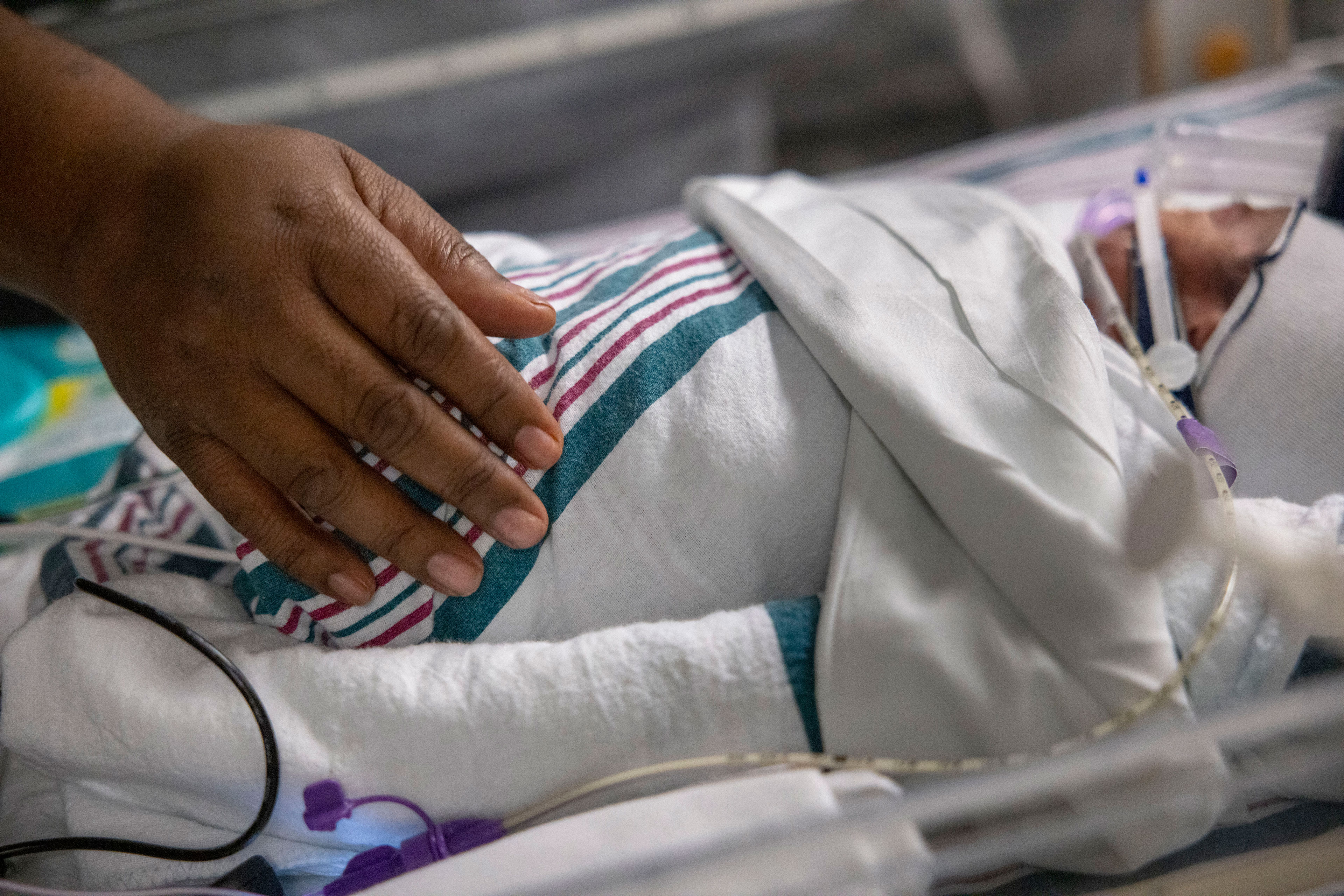 A close-up photo of a hand on a swaddled baby in an ICU bed.