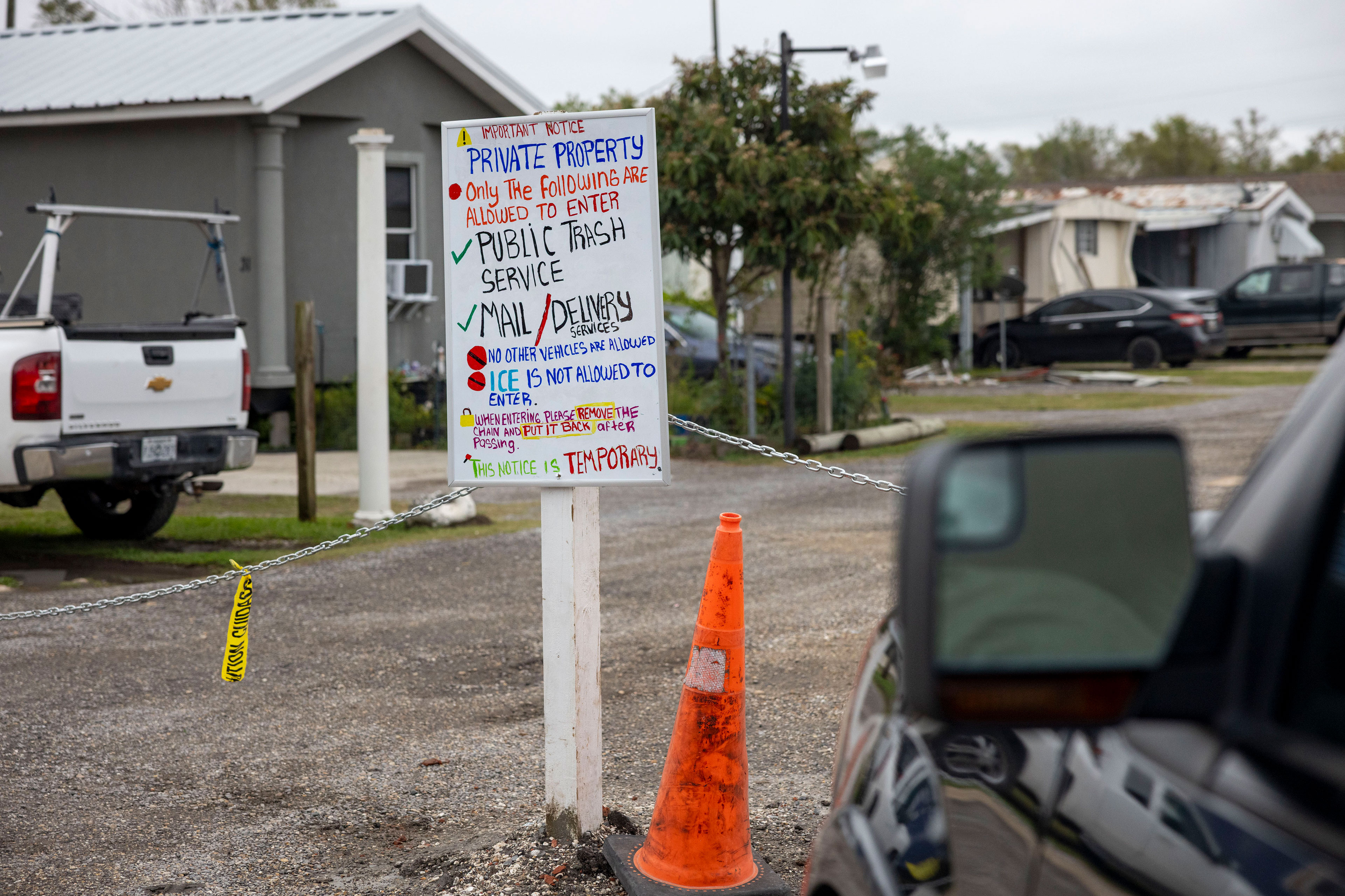 A sign is posted in a road that has a chain stretched across it. The sign reads: "Important Notice, Private Property, Only the following are allowed to enter: Public trash service, mail/delivery services. No other vehicles are allowed. ICE is not allowed to enter. When entering please remove the chain and put it back after passing. This notice is temporary."