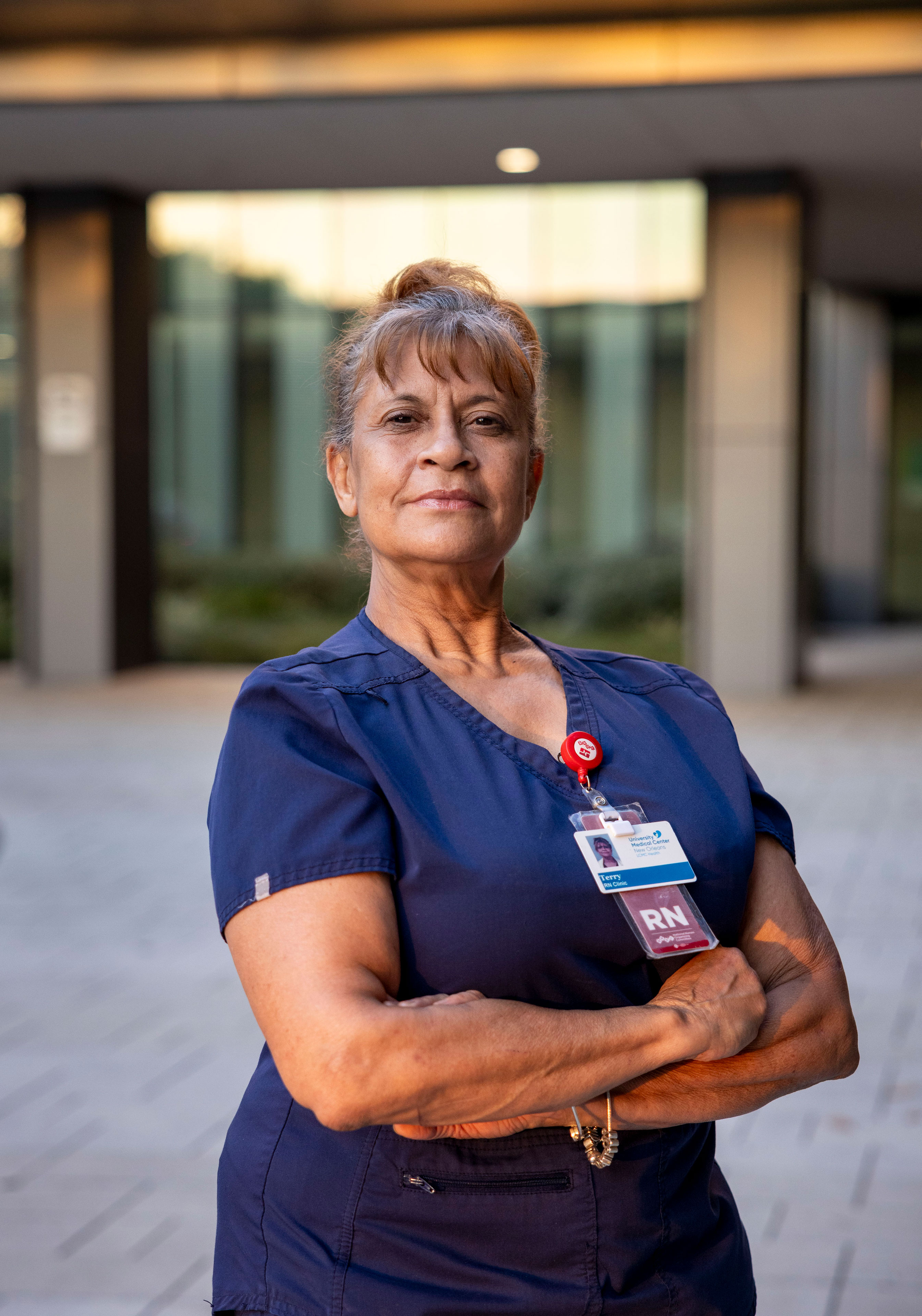 A woman in dark blue scrubs stands outside a building at sunset and looks at the camera.