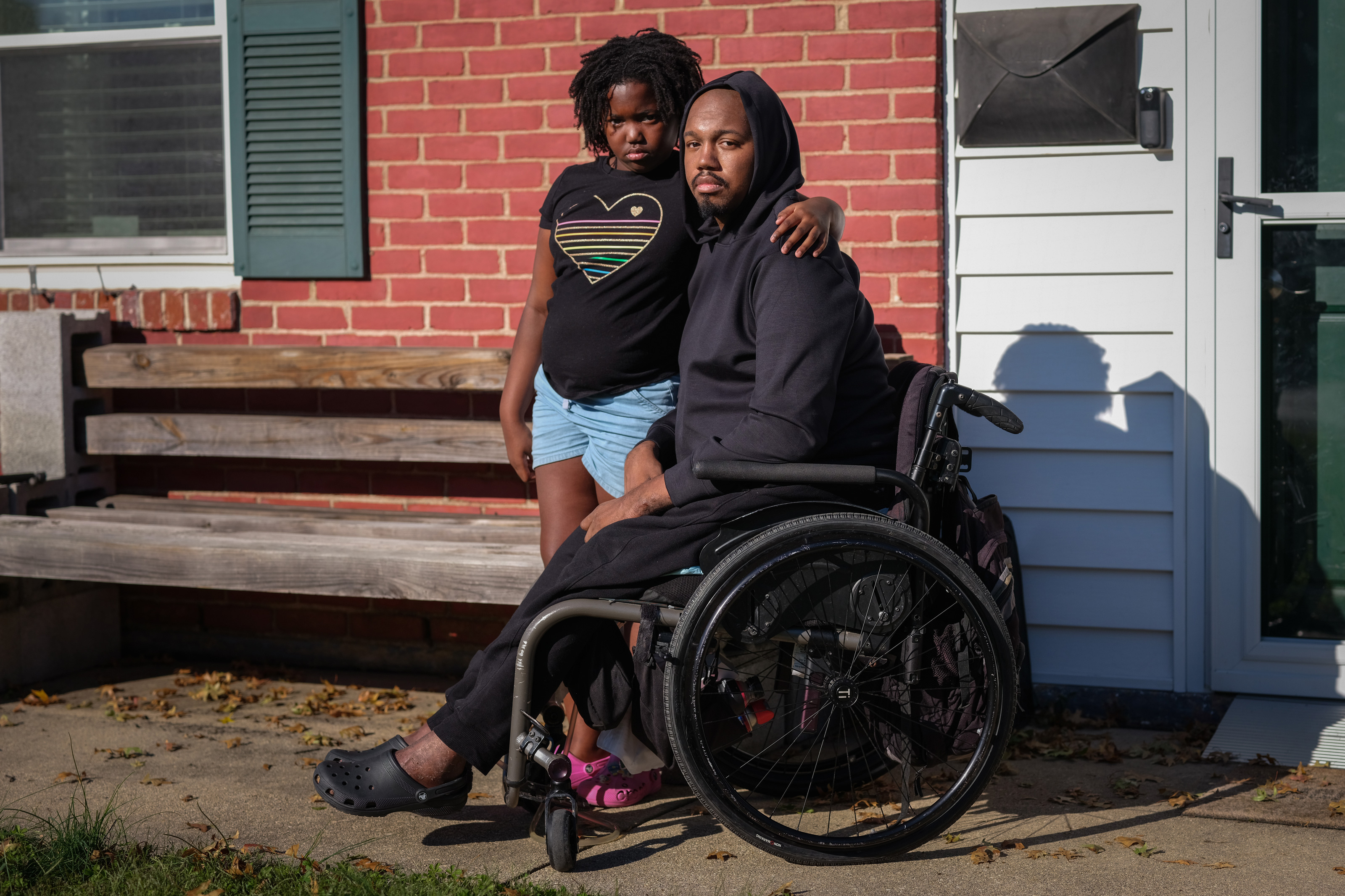 A photo of a Black man seated in a wheelchair looks at the camera. Behind him is his young daughter.