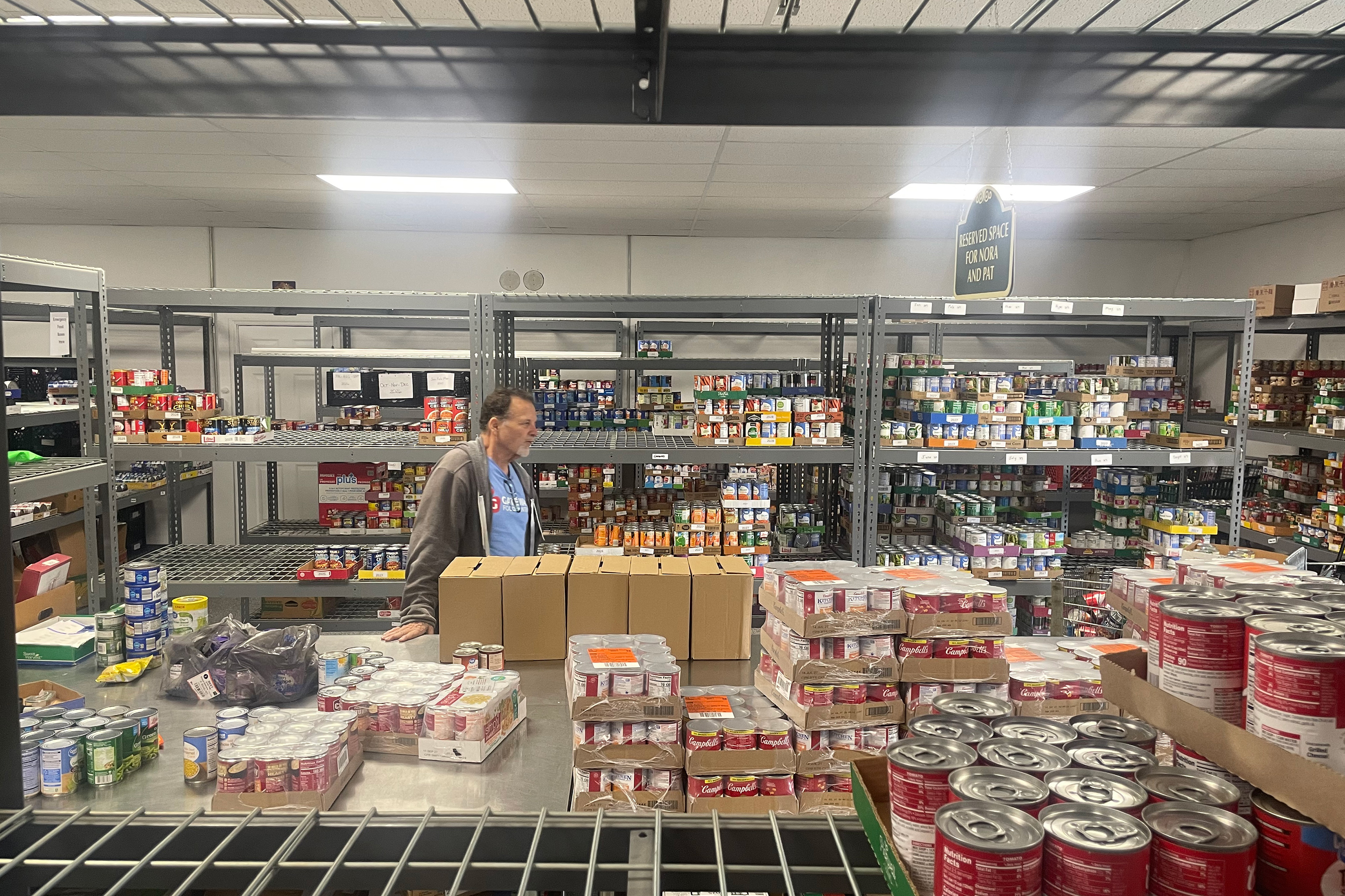 A man stands in a food pantry with shelves and tables of on non-perishable goods surrounding him