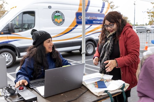 A photo of two women in cold weather wear. The woman on the left has a laptop and keyboard in front of her. The woman on the right is speaking to her. Paperwork and pamphlets are on the table in front of them.
